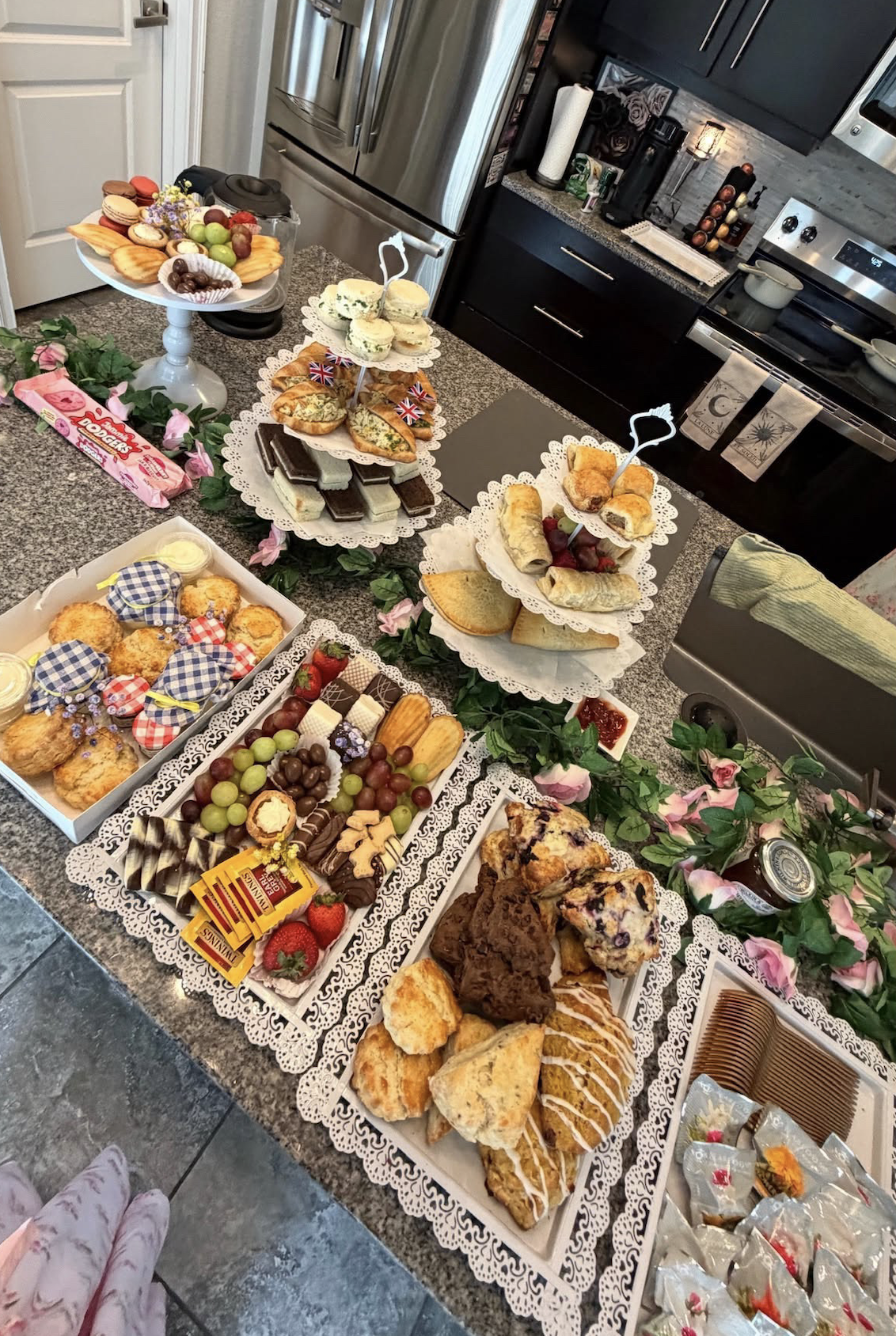 A table filled with various desserts and snacks, including cookies, cakes, sandwiches, pastries, chocolates, and fresh fruit, decorated with pink flowers and greenery.