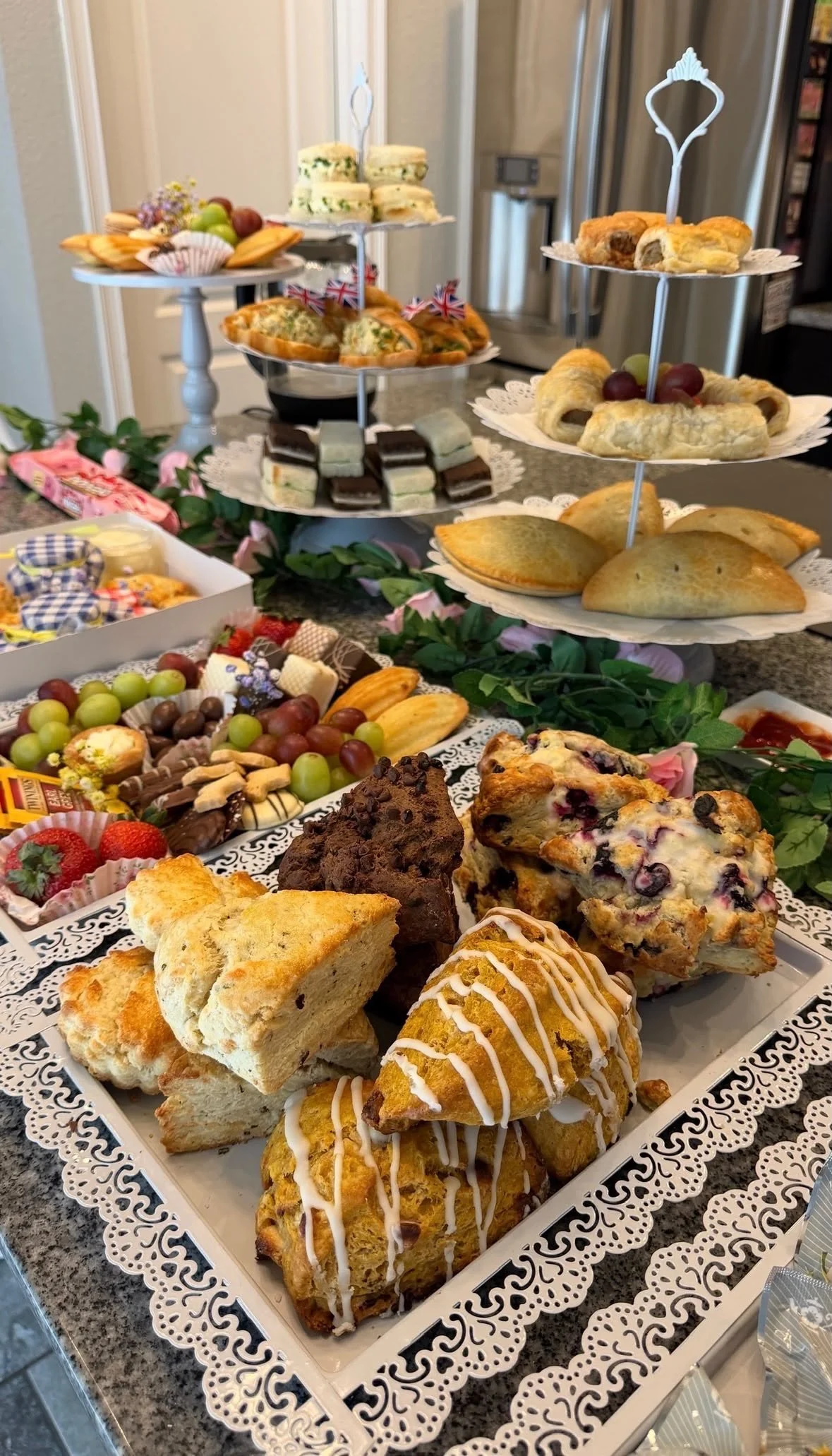 A tiered tray of various baked goods including scones, biscuits, and muffins, surrounded by fruit and candies, set on a decorated table.
