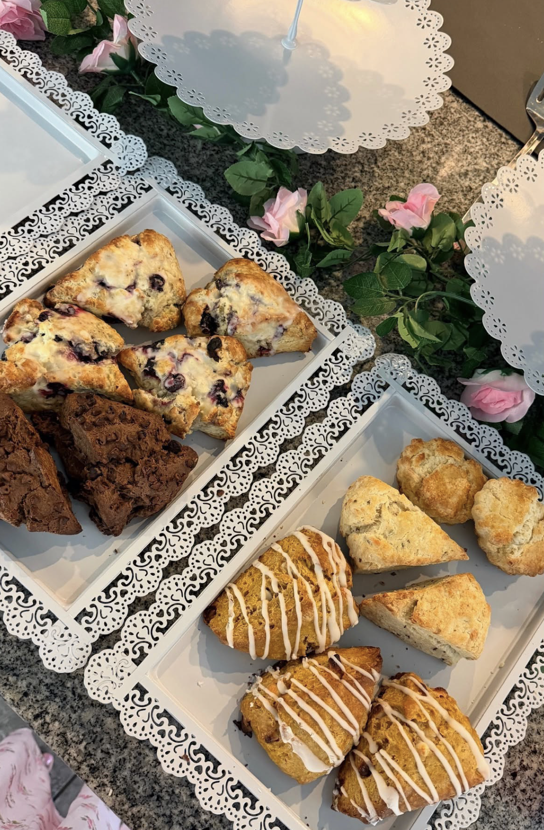 Assorted baked goods on white lace doilies, including blueberry scones, chocolate chip loaf, and bread slices, decorated with pink roses around the display.
