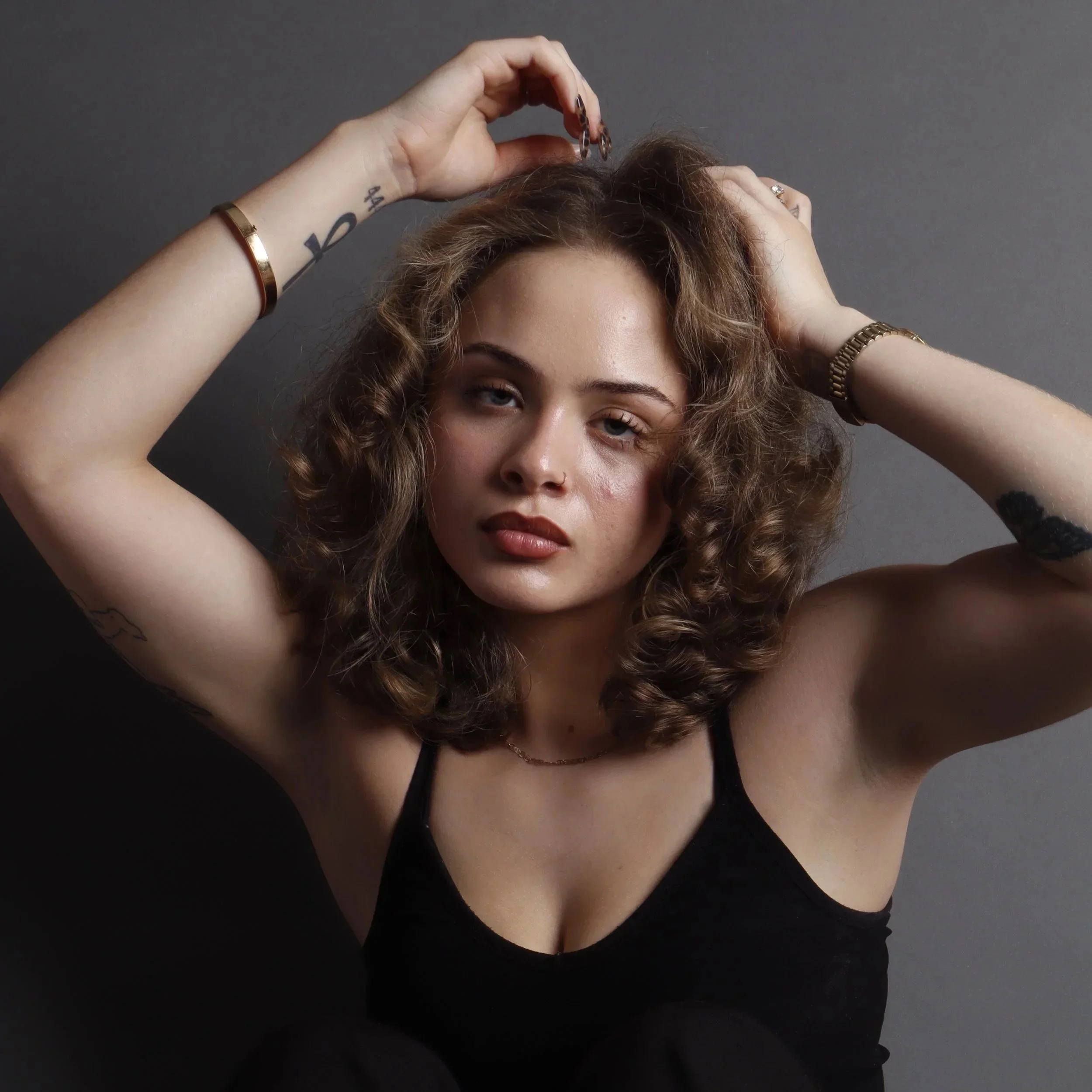 A woman with curly hair, wearing multiple rings and gold bracelets, posing with her hands in her hair against a dark background.
