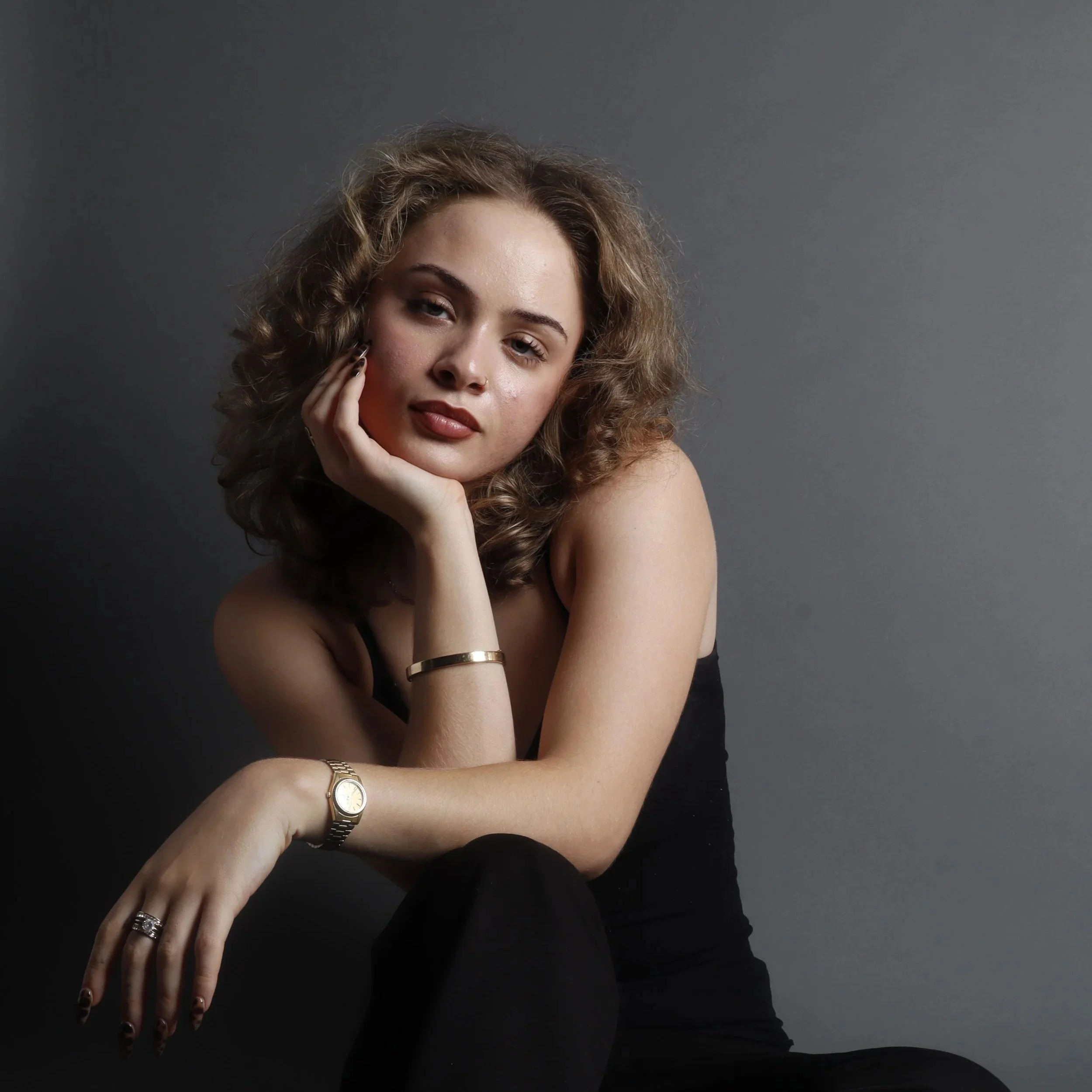 A woman with curly hair wears a black top, gold jewelry, and has a neutral facial expression, resting her hand on her face, against a dark gray background.