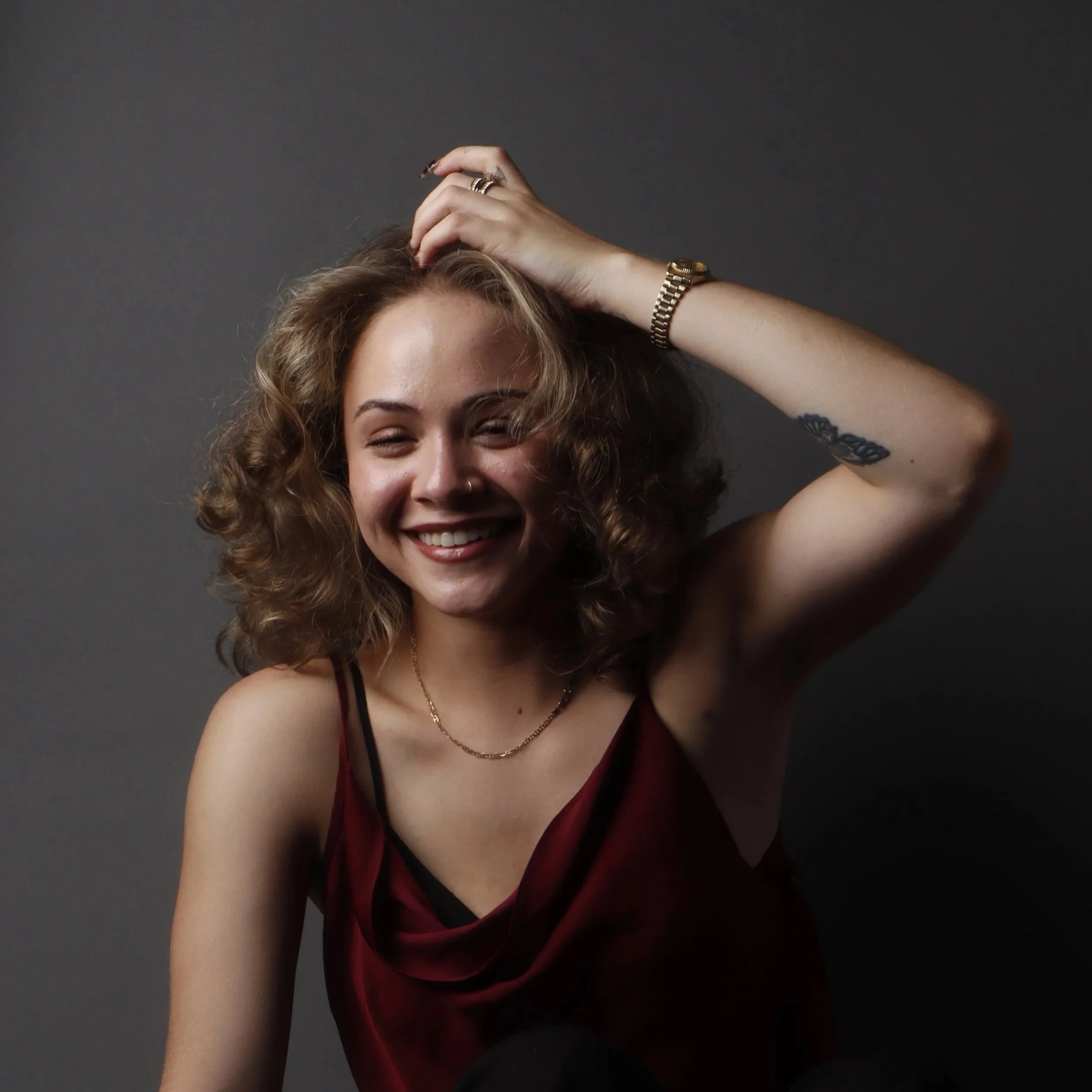 A young woman with curly hair and a nose ring smiling, wearing a sleeveless top, gold watch, and rings, against a dark gray background.