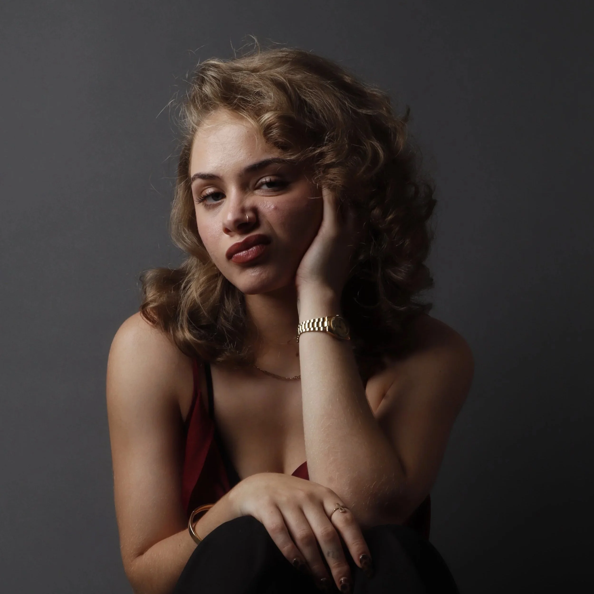 A young woman with curly hair, a nose piercing, and dark lipstick, holding her face with her hand, wearing gold jewelry, against a gray background.