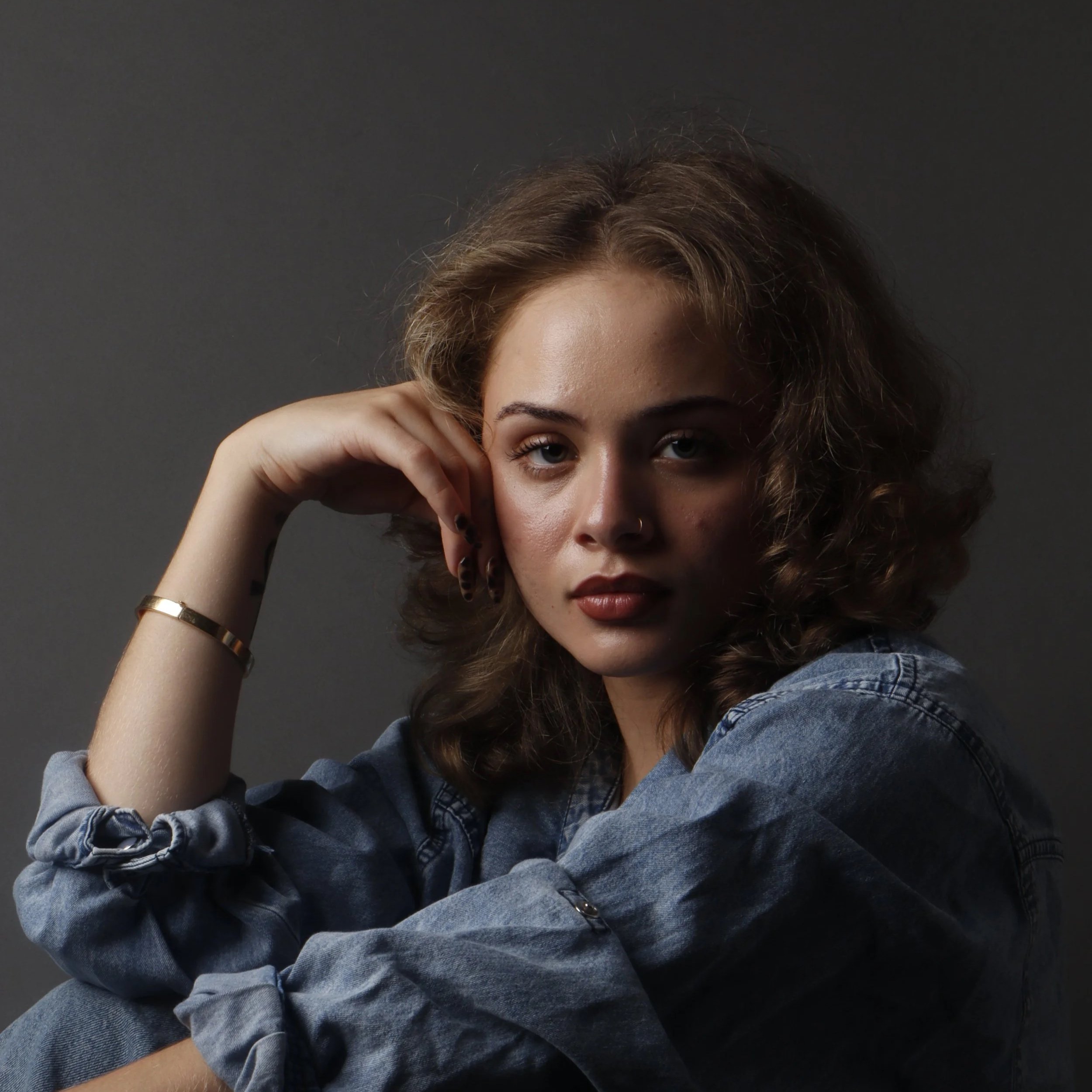 Close-up of a woman with curly hair, wearing a denim jacket, and touching her face with her right hand, against a dark background.
