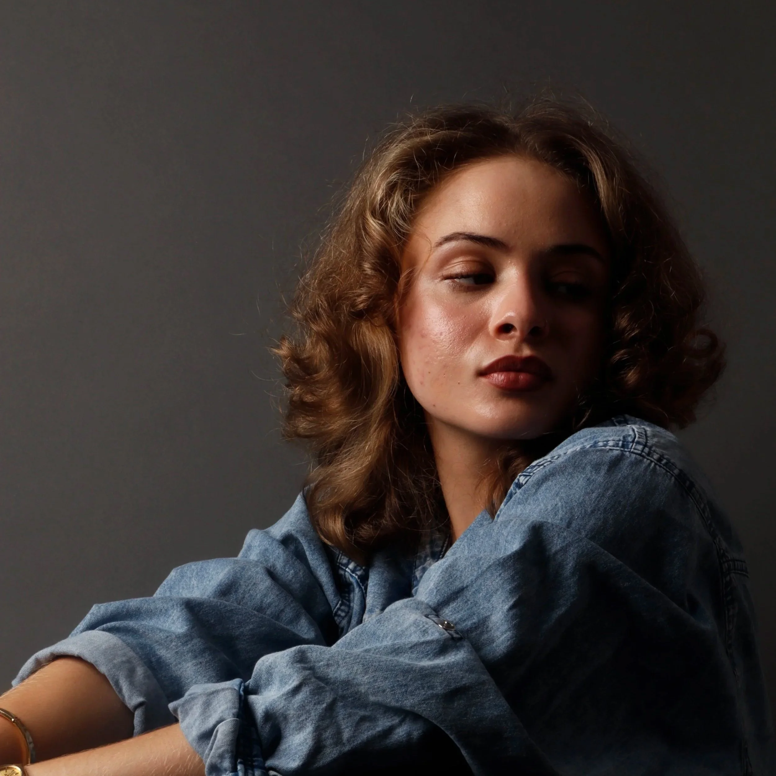 Close-up of a woman with curly hair wearing a denim jacket, standing against a dark gray background.
