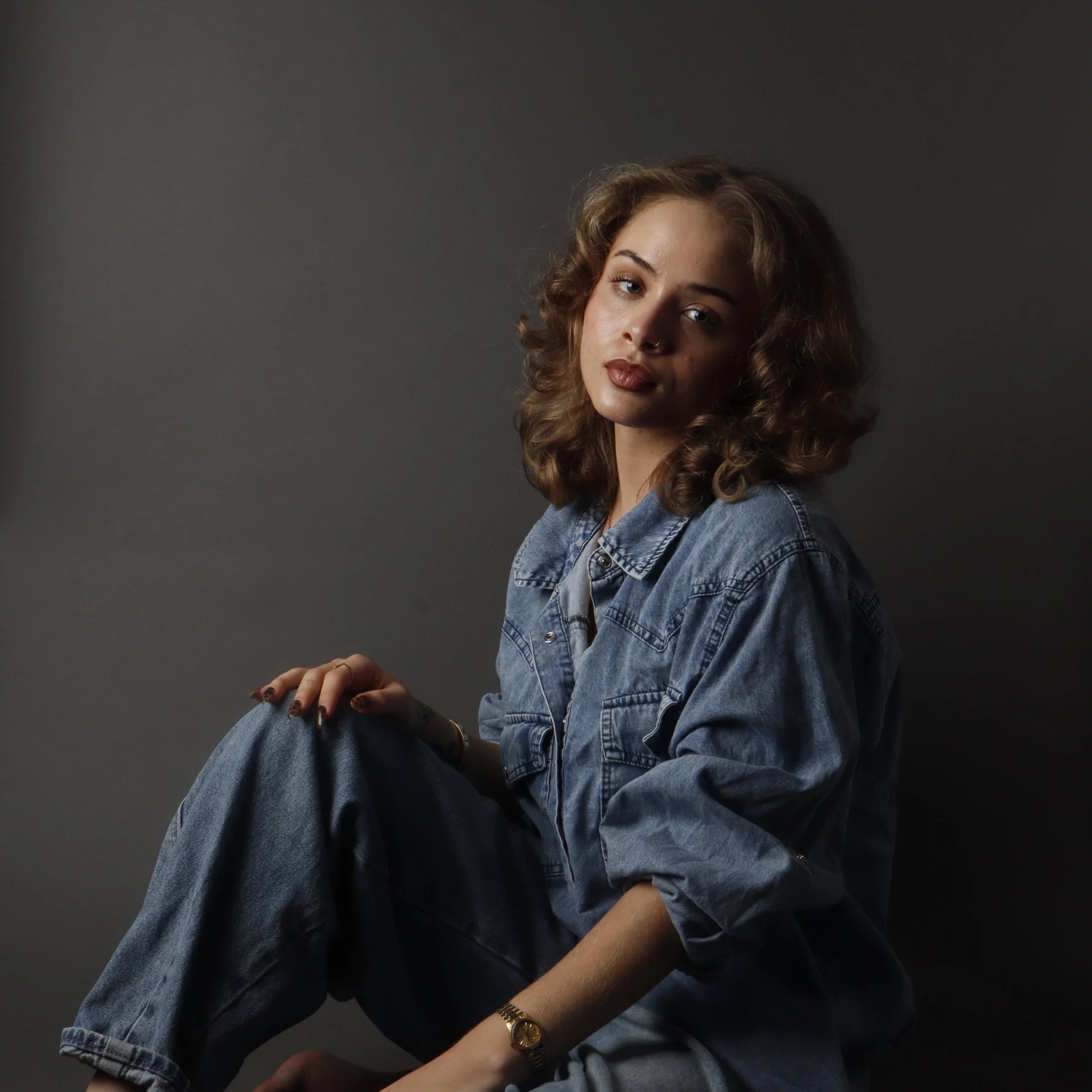 A young woman with curly brown hair and blue eyes wearing a denim jacket, with her hand on her knee and a gold watch on her wrist, is sitting against a gray background.