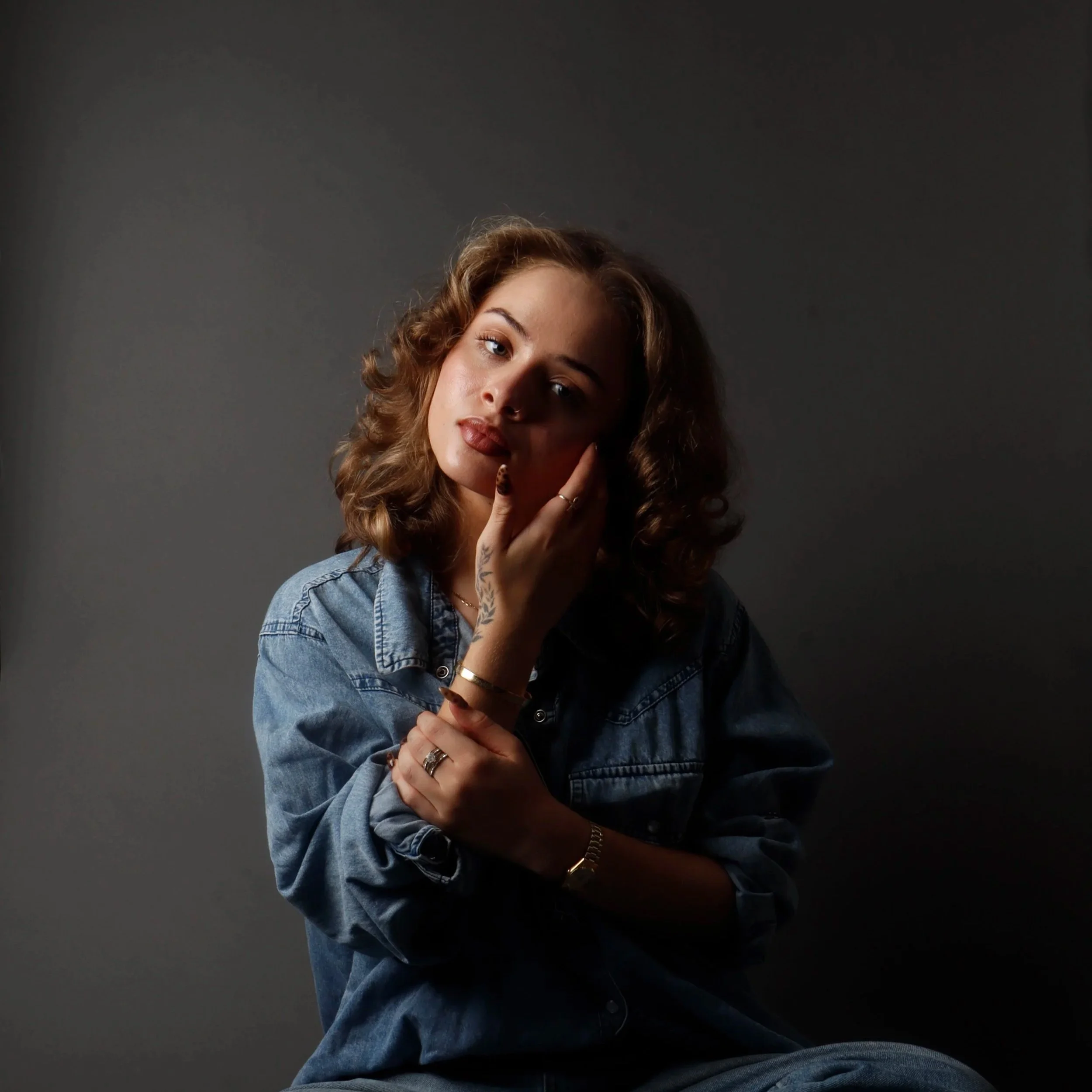 A woman with curly hair and wearing a denim shirt poses against a dark gray background.