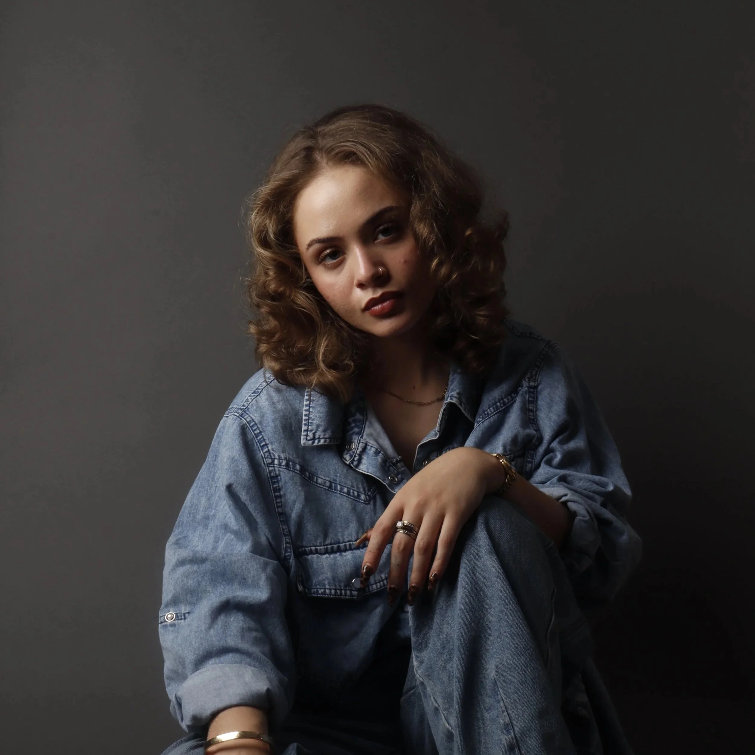 A young woman with curly hair wearing a denim jacket and jewelry, posing against a dark background.