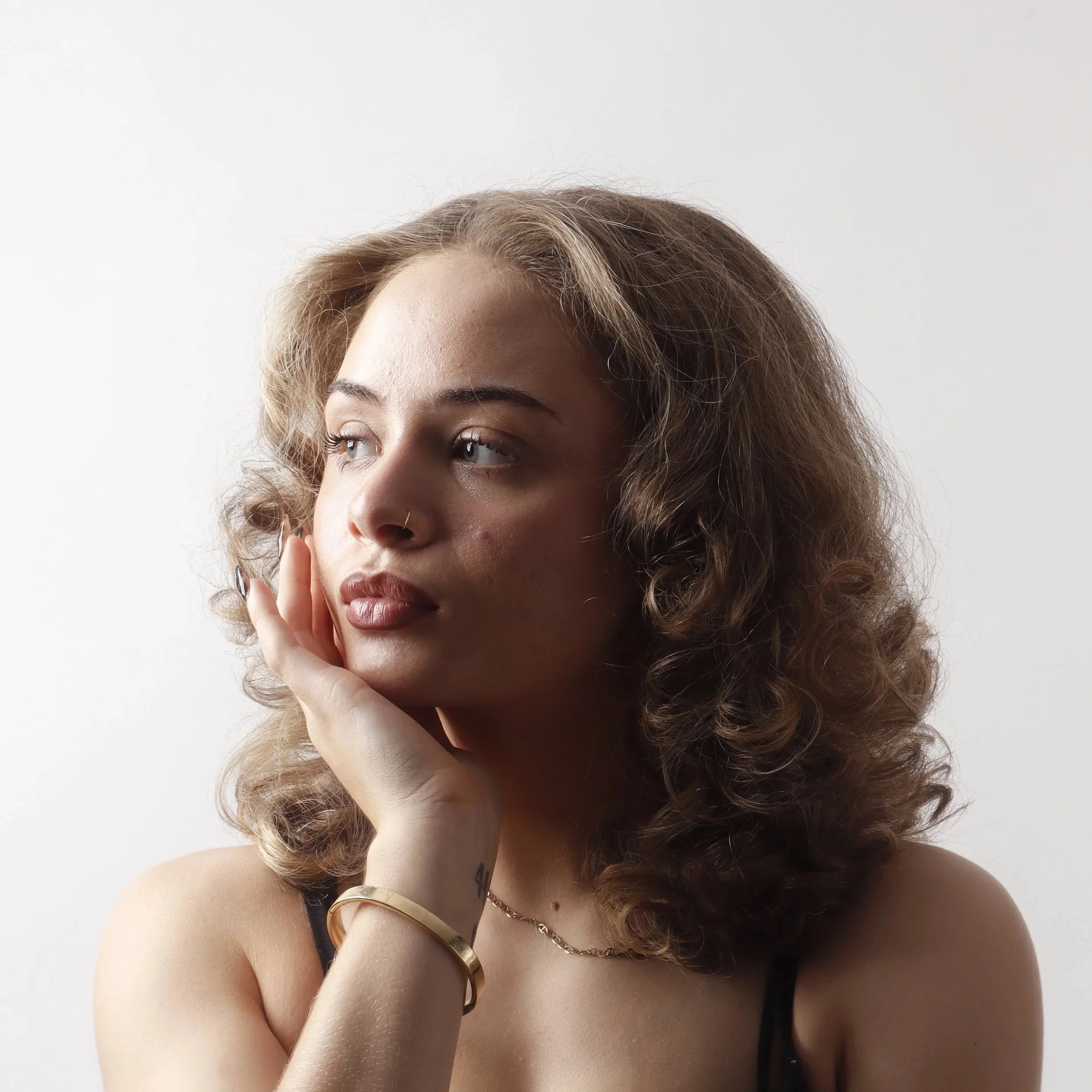 A woman with curly brown hair is resting her chin on her hand, wearing a gold bracelet and a thin chain necklace, with a neutral background.
