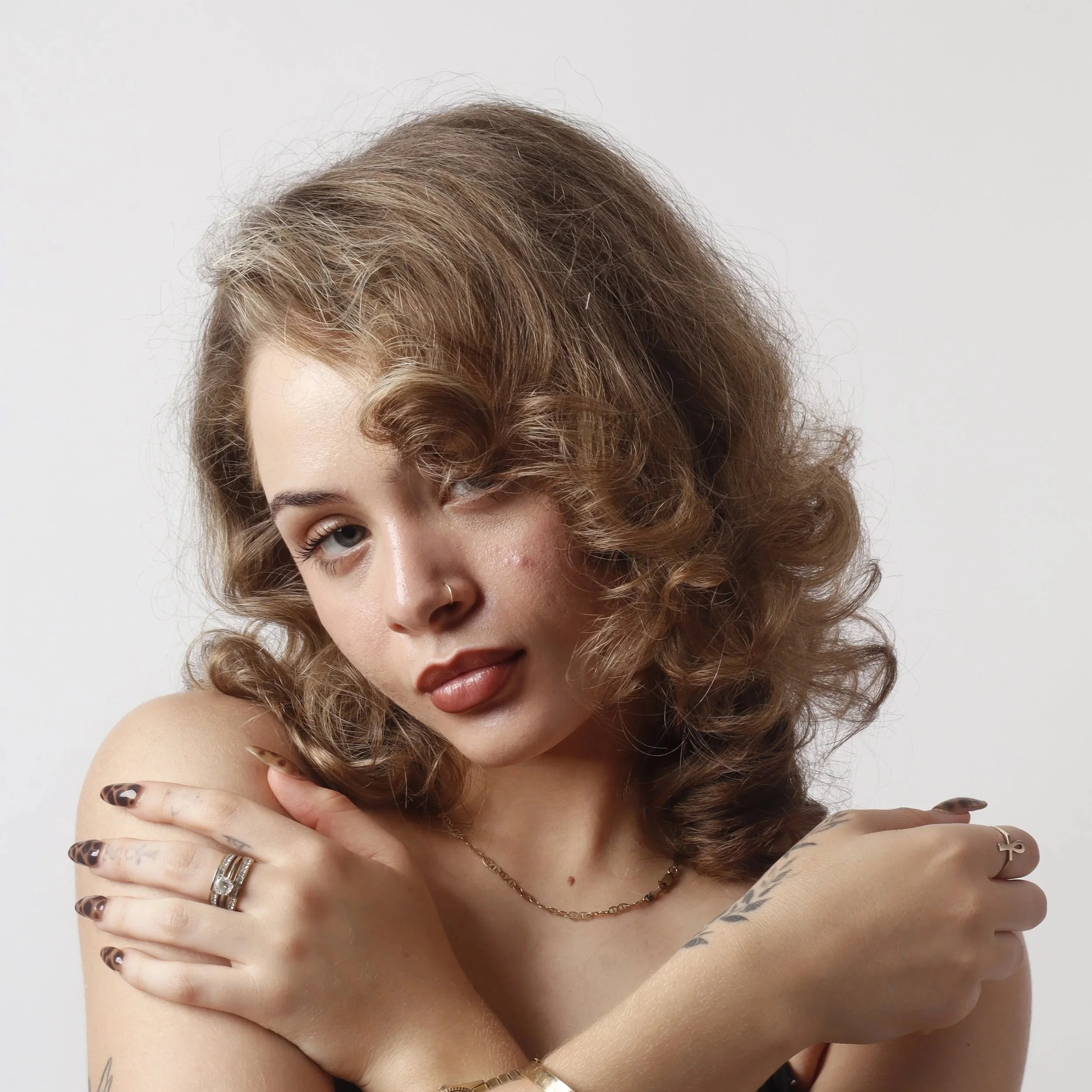 A person with curly, light brown hair, which is styled voluminously and slightly tousled, covering part of their face. They are topless, with their arms crossed over their chest, and displaying multiple rings on their fingers, along with a thin neckl