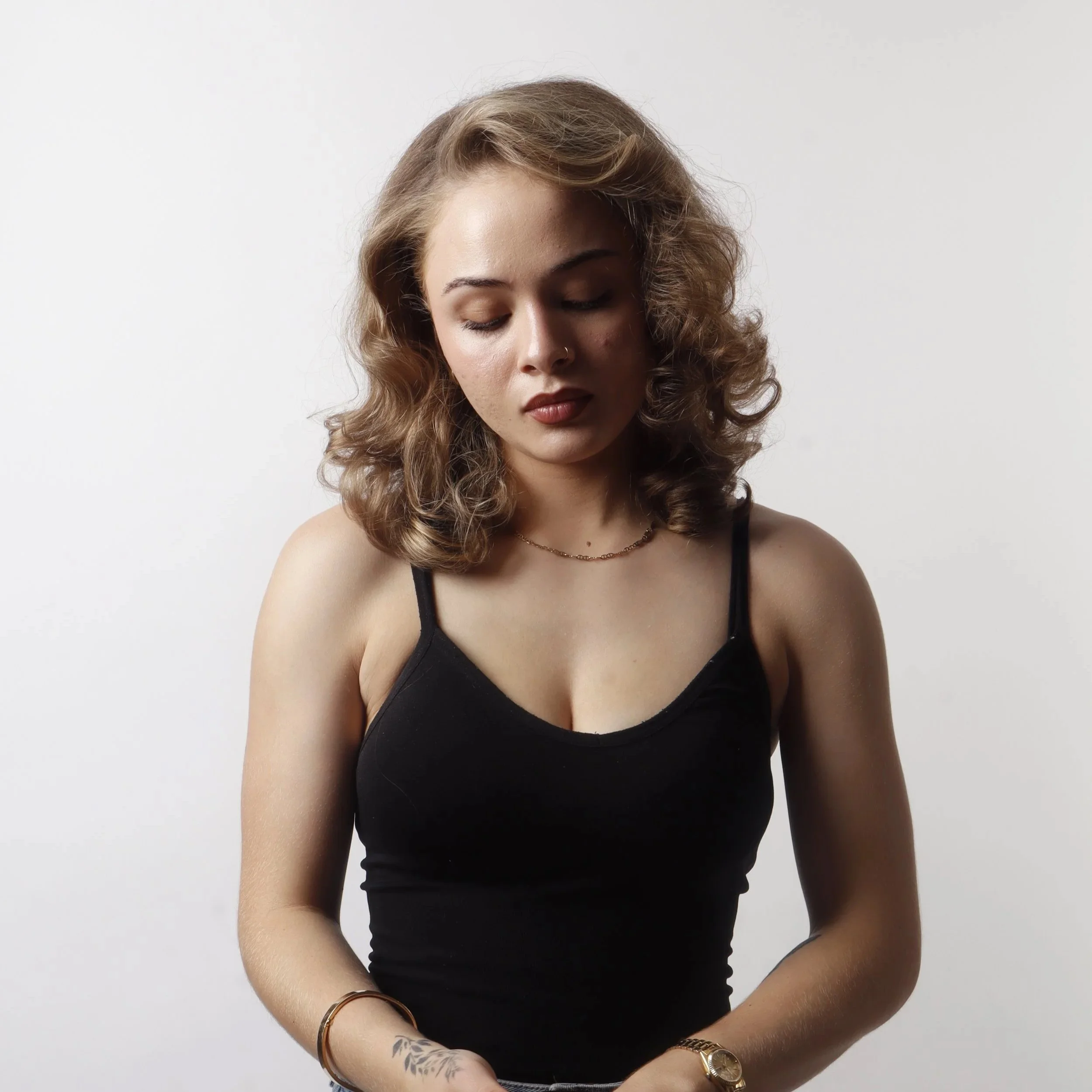A woman with wavy light brown hair, closed eyes, and nose piercings wearing a black spaghetti strap top and gold jewelry, standing against a white background.