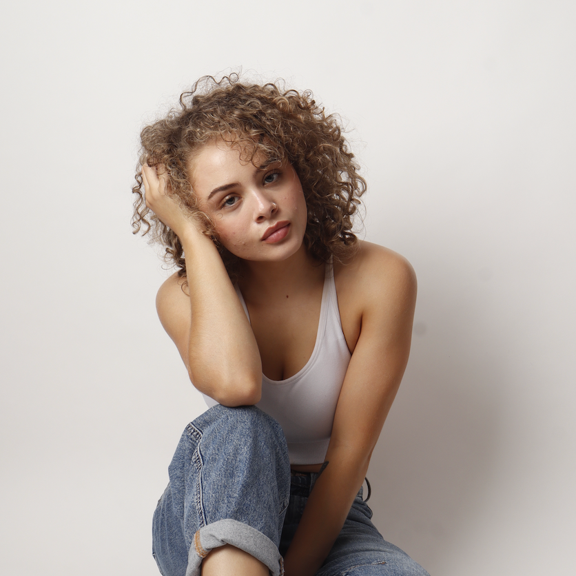 Young woman with curly hair sitting against a white wall, wearing a white tank top and jeans, with a relaxed and thoughtful expression.