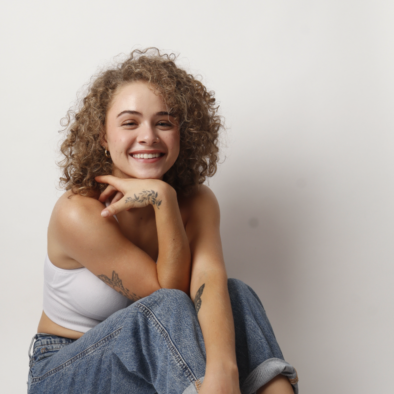 Young woman with curly hair smiling, sitting with her arms around her knees against a plain white background.