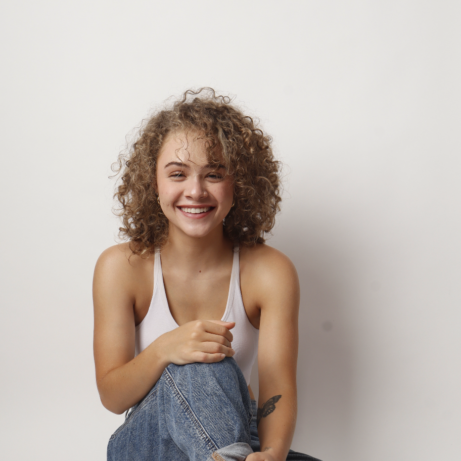 A young woman with curly hair smiling, wearing a white tank top and jeans, sitting against a plain white background.