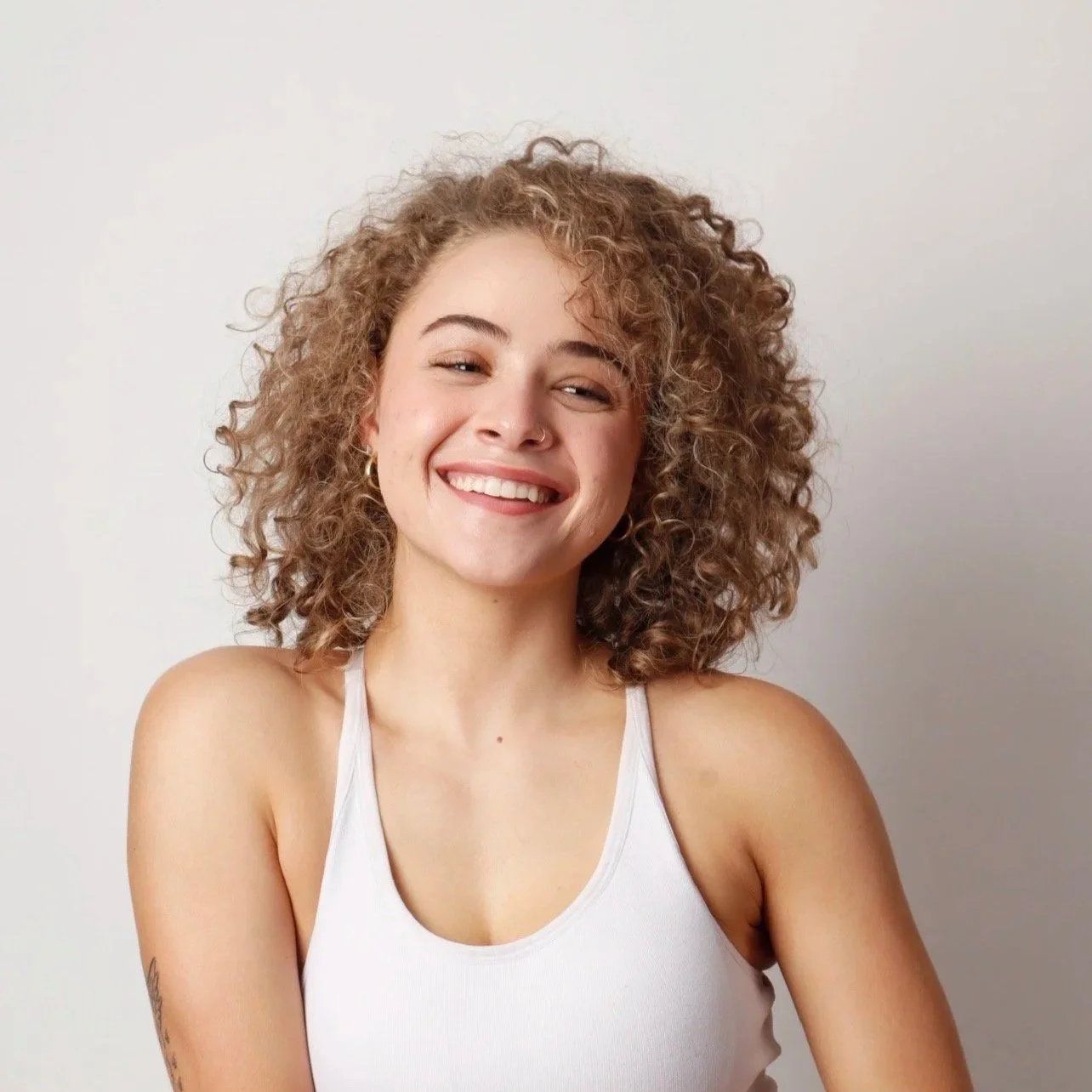 A young woman with curly hair smiling and wearing a white tank top against a plain light background.