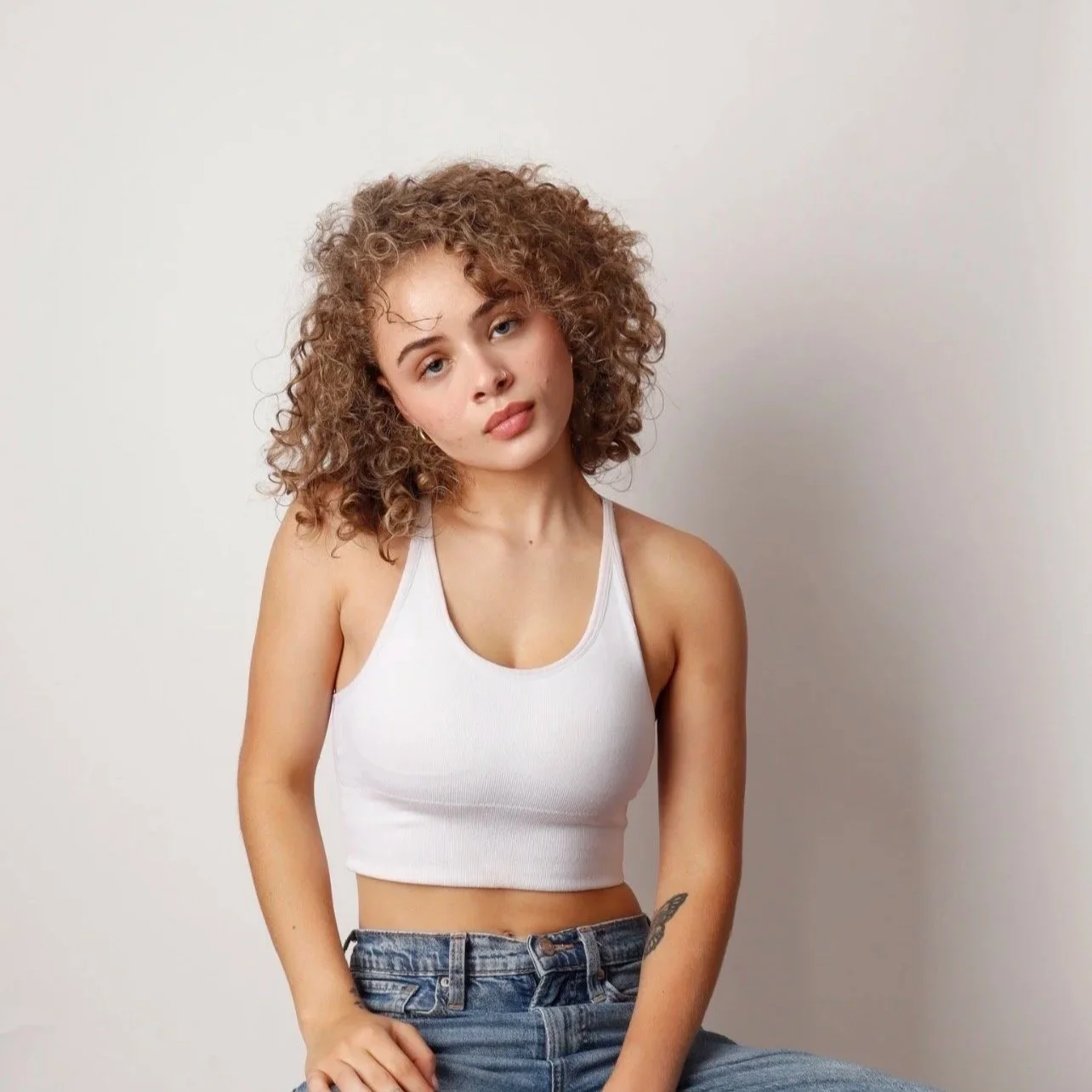 A young woman with curly hair wearing a white tank top and denim jeans, sitting against a plain light background.