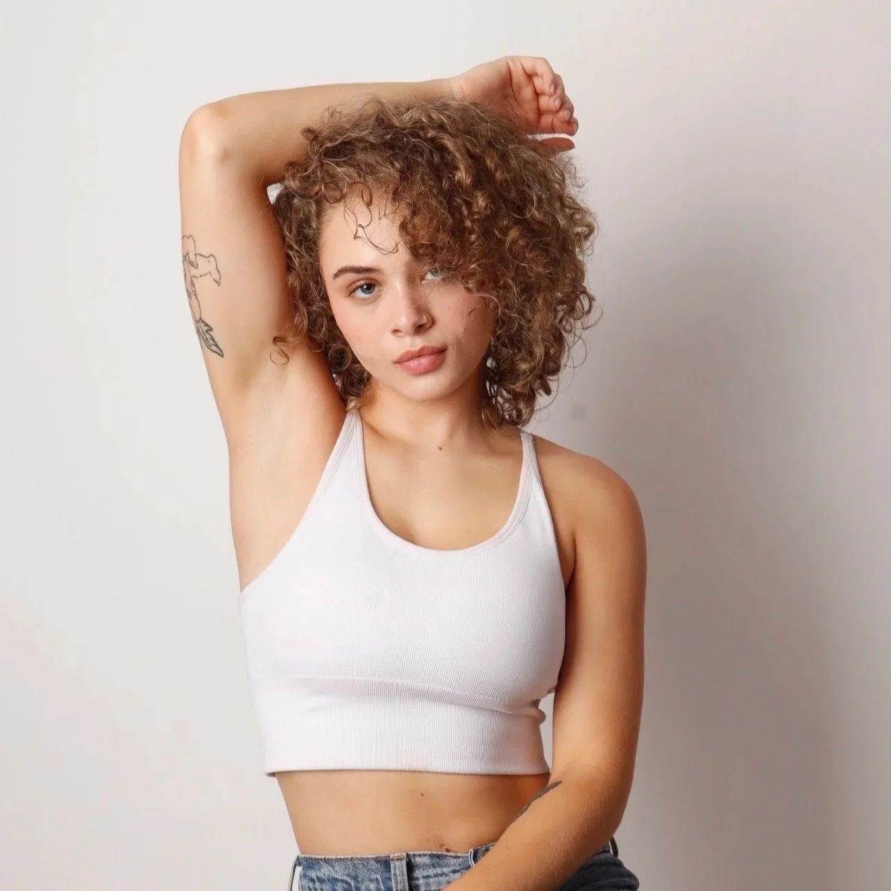 A young woman with curly hair, wearing a white tank top, is posing with her right arm raised above her head and her left arm down, against a plain white background.