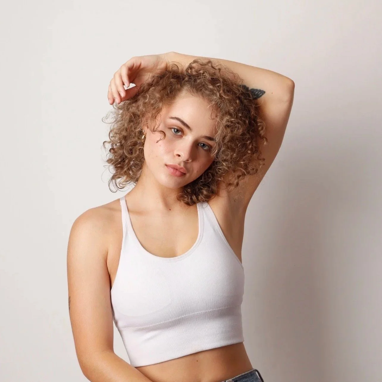 A young woman with curly hair and light skin posing with one arm raised above her head, wearing a white tank top and looking at the camera.