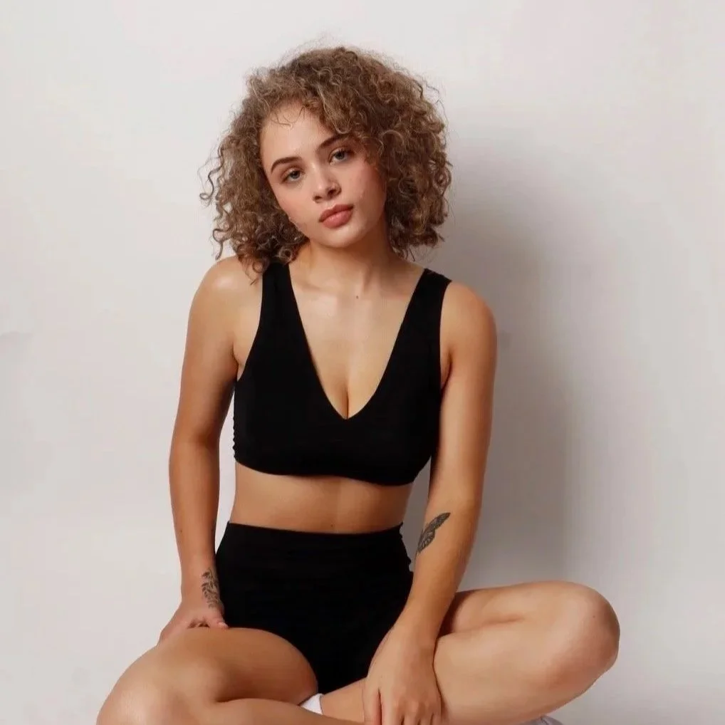 Young woman with curly hair wearing a black crop top and black shorts, sitting cross-legged against a plain light-colored wall.