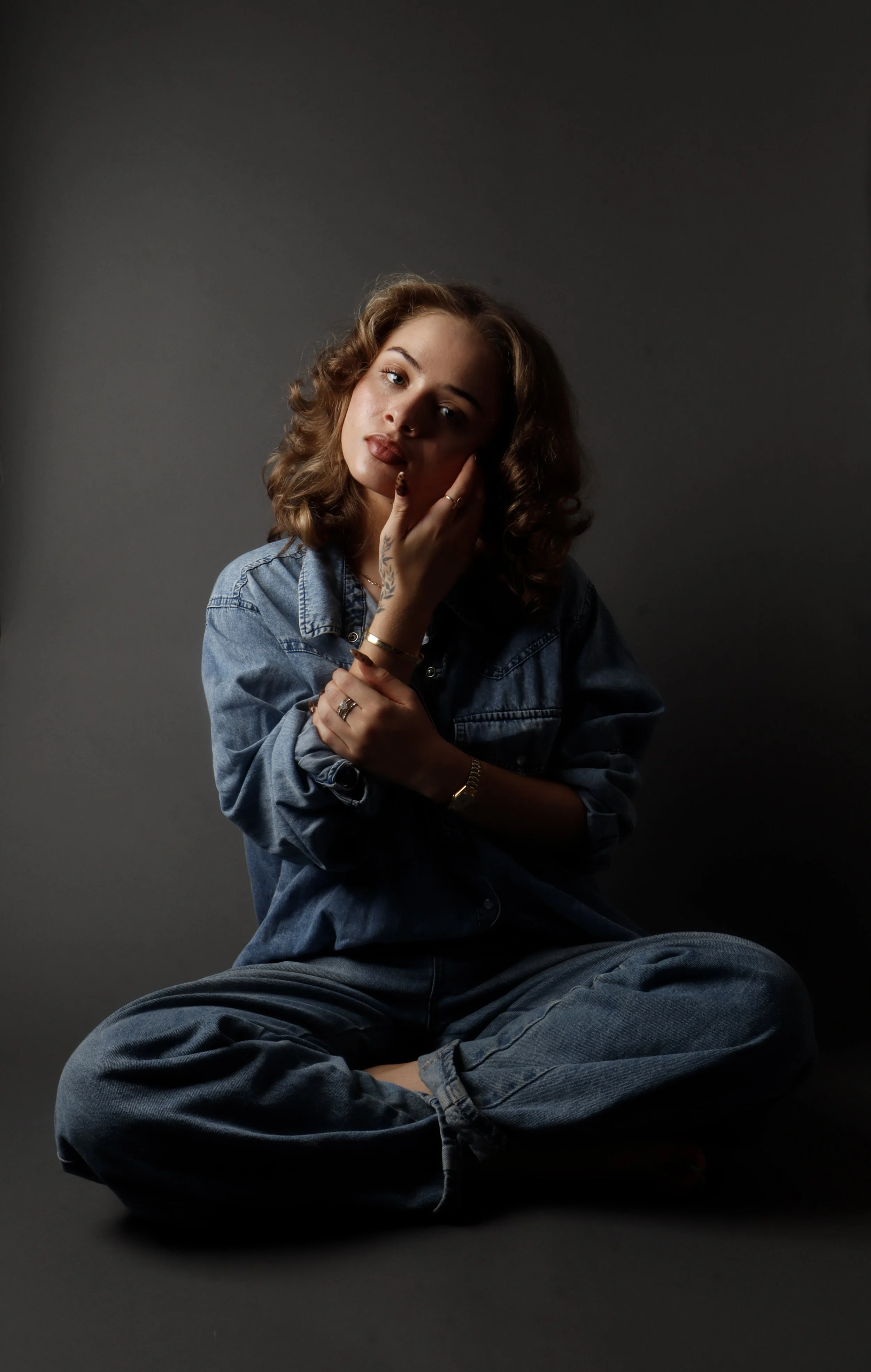 A young woman with curly brown hair sits cross-legged on a dark surface against a dark background, wearing a denim jacket and jeans, with her hand touching her cheek and looking directly at the camera.