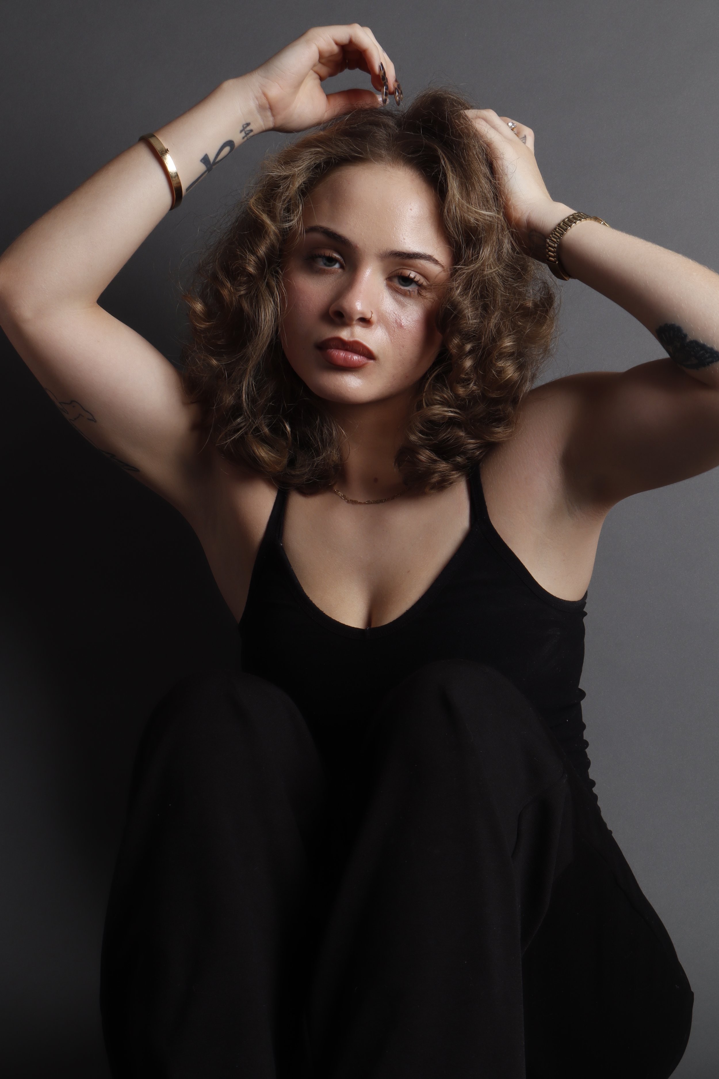 A woman with curly hair wearing a black top, sitting against a dark background, with her arms raised and hands on her head.