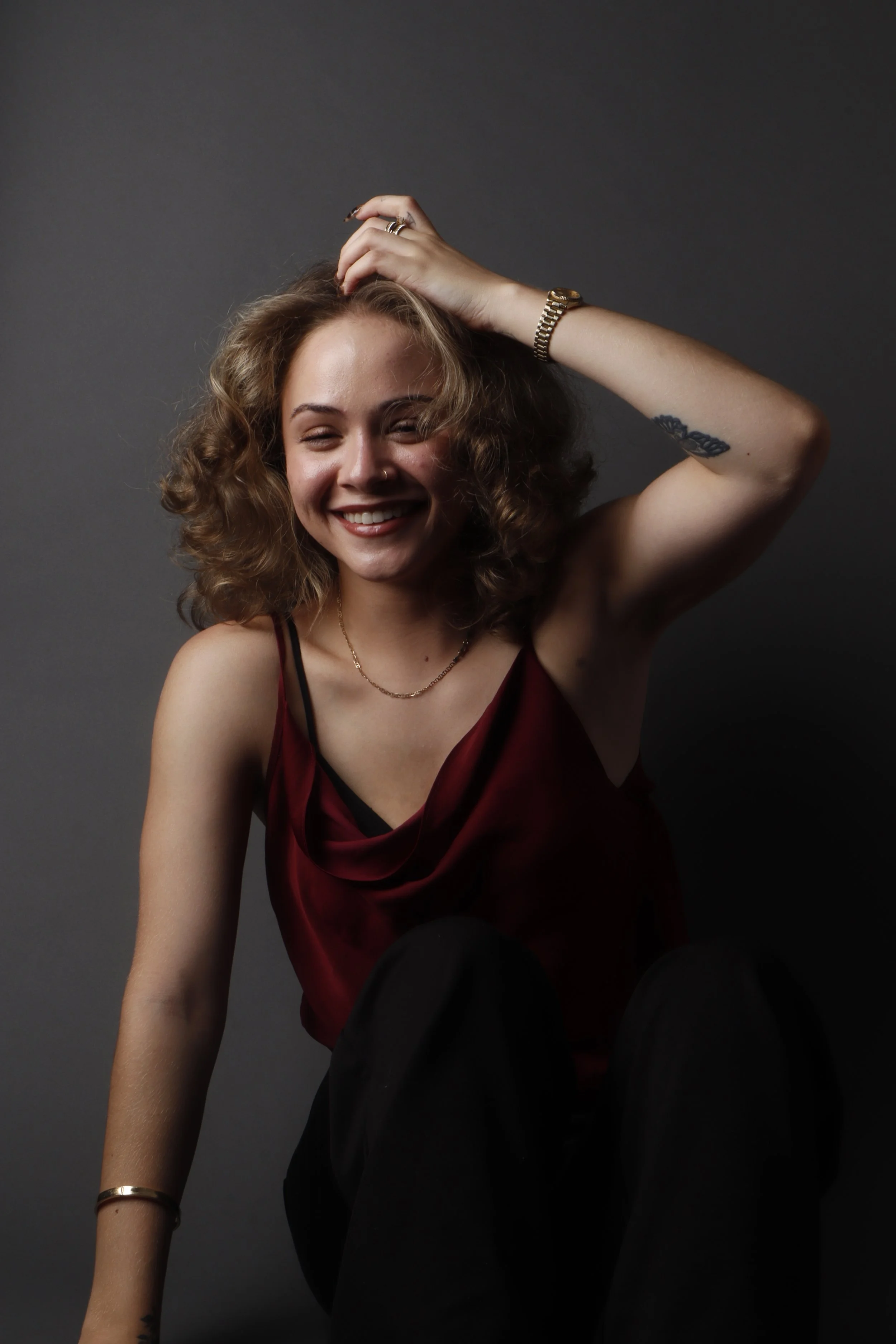 A young woman with curly hair smiling, wearing a maroon satin top, gold jewelry, and black pants against a dark gray background.