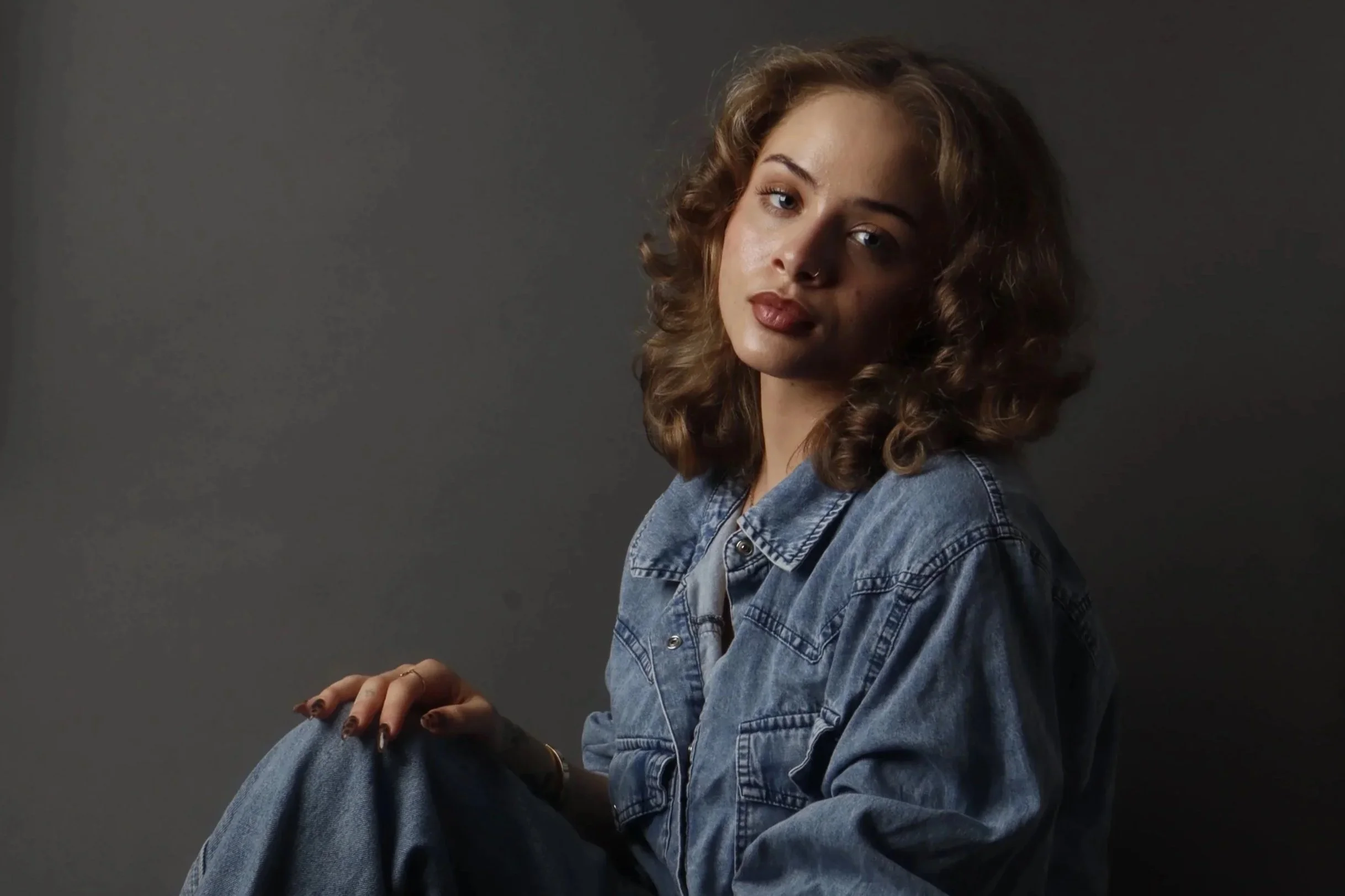 Portrait of a woman with curly hair wearing a denim jacket, sitting against a dark gray background.