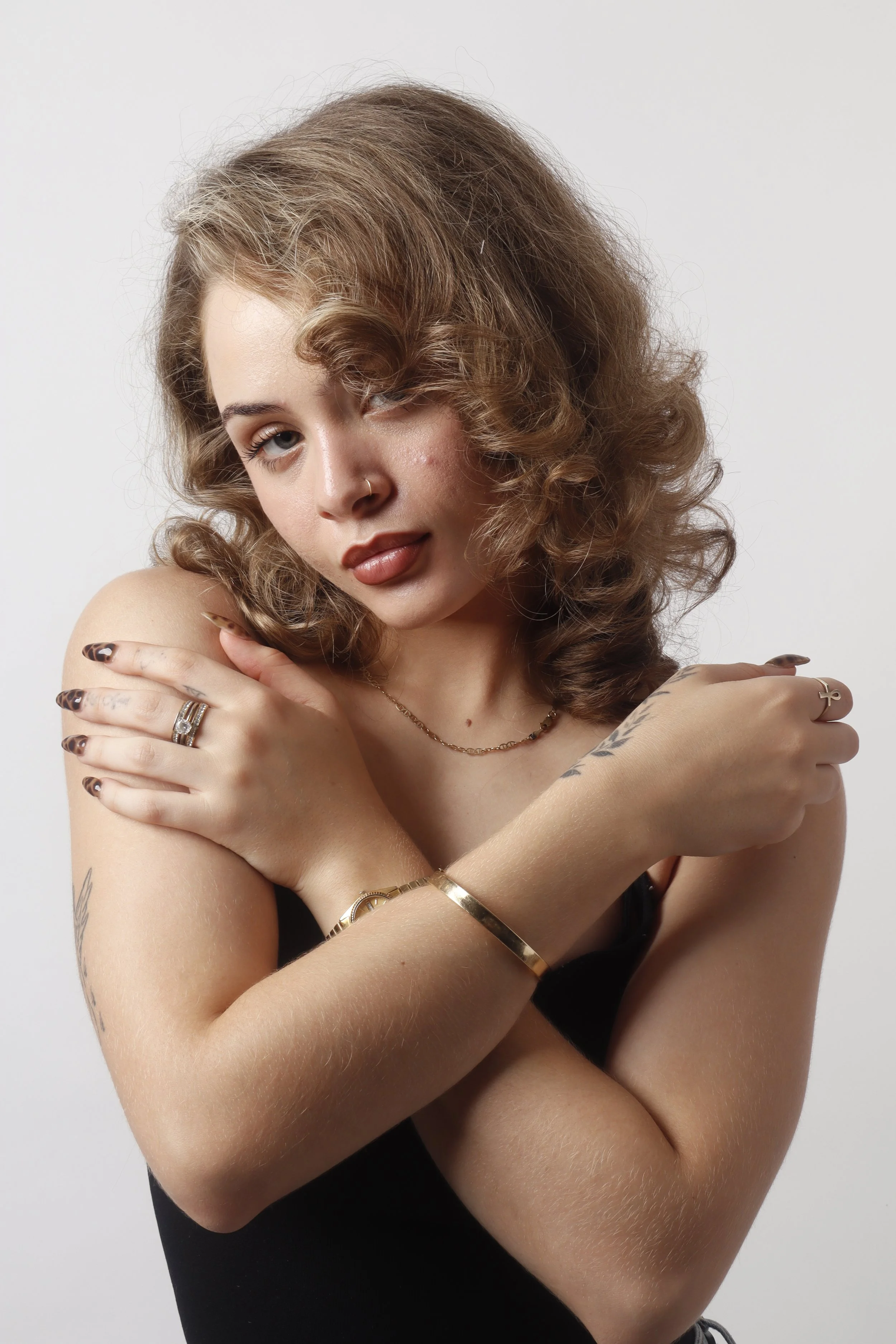 A woman with curly hair and tattoos hugging herself against a plain white background.