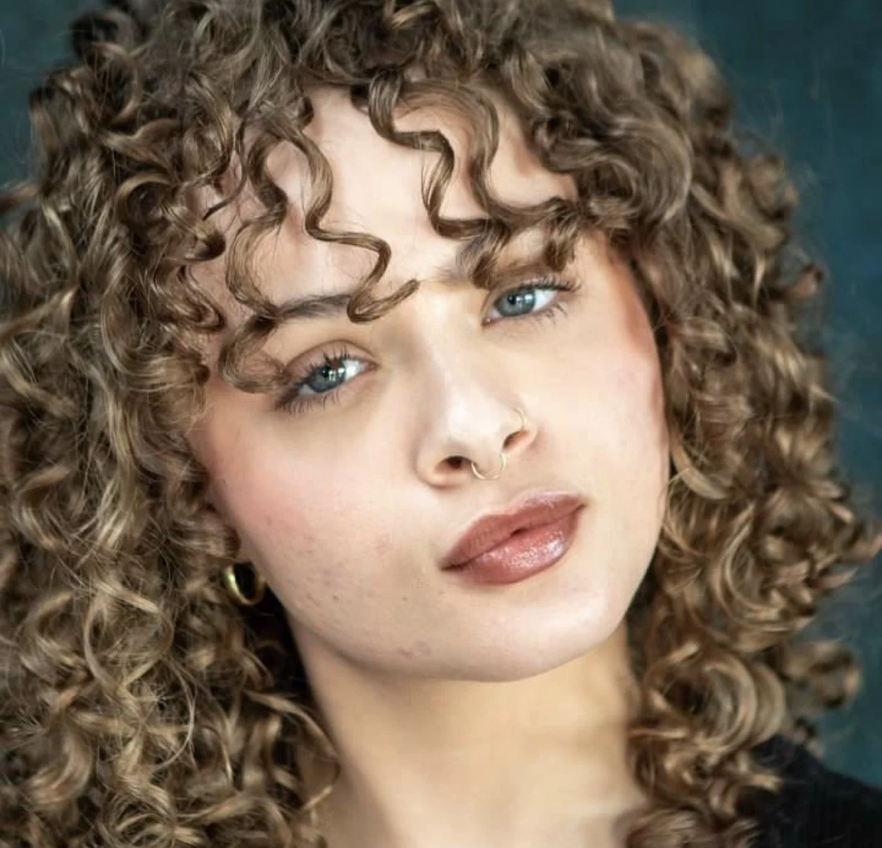 A woman with blue eyes, curly brown hair, and a septum piercing looks at the camera.