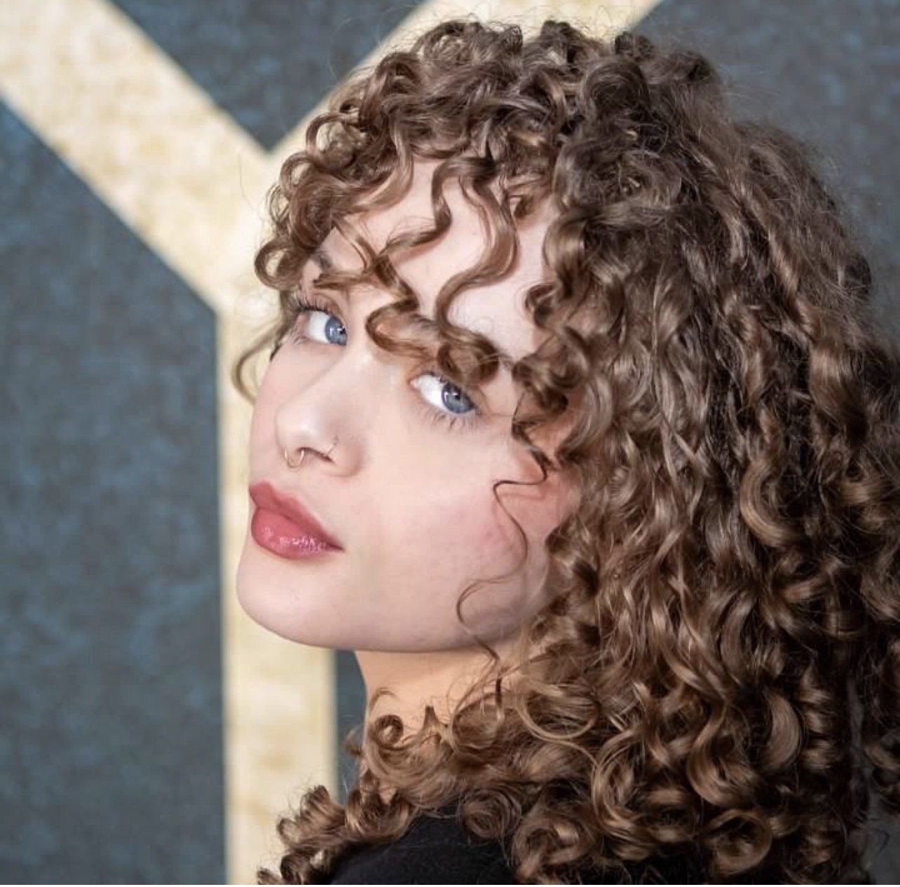 Close-up of a woman with curly brown hair, blue eyes, and a septum piercing, looking over her shoulder.
