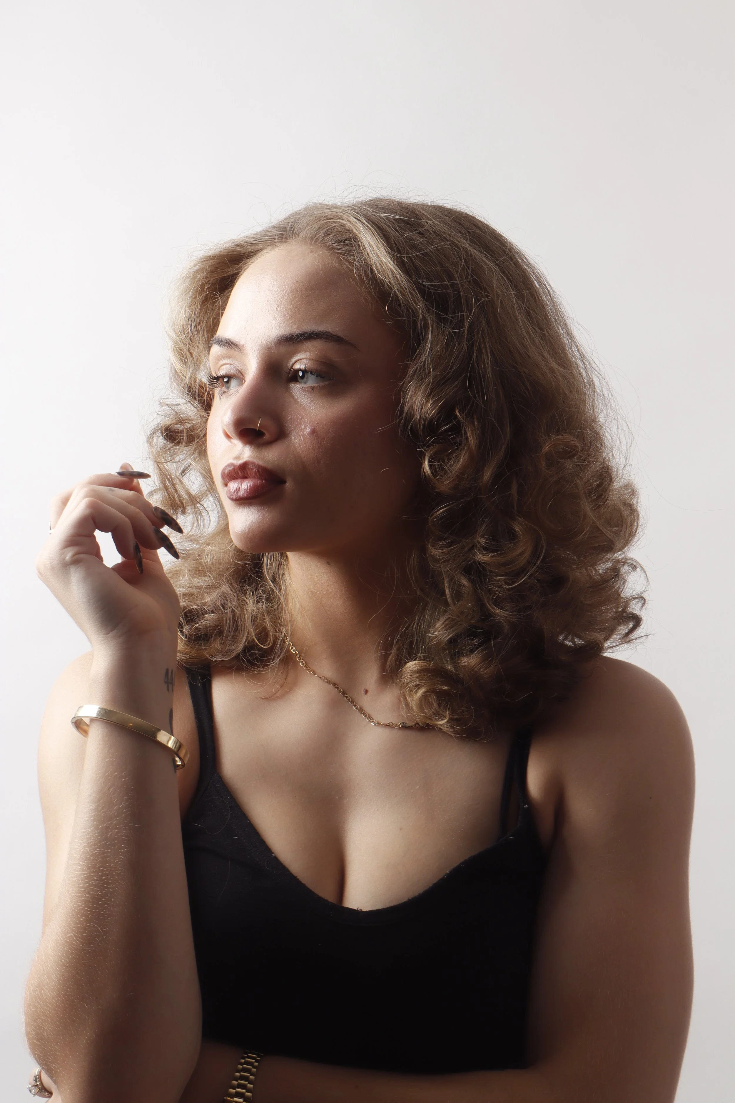 A woman with curly, shoulder-length hair, wearing a black tank top, gold jewelry, and a nose ring, looking to her left with her hand near her face, against a plain white background.