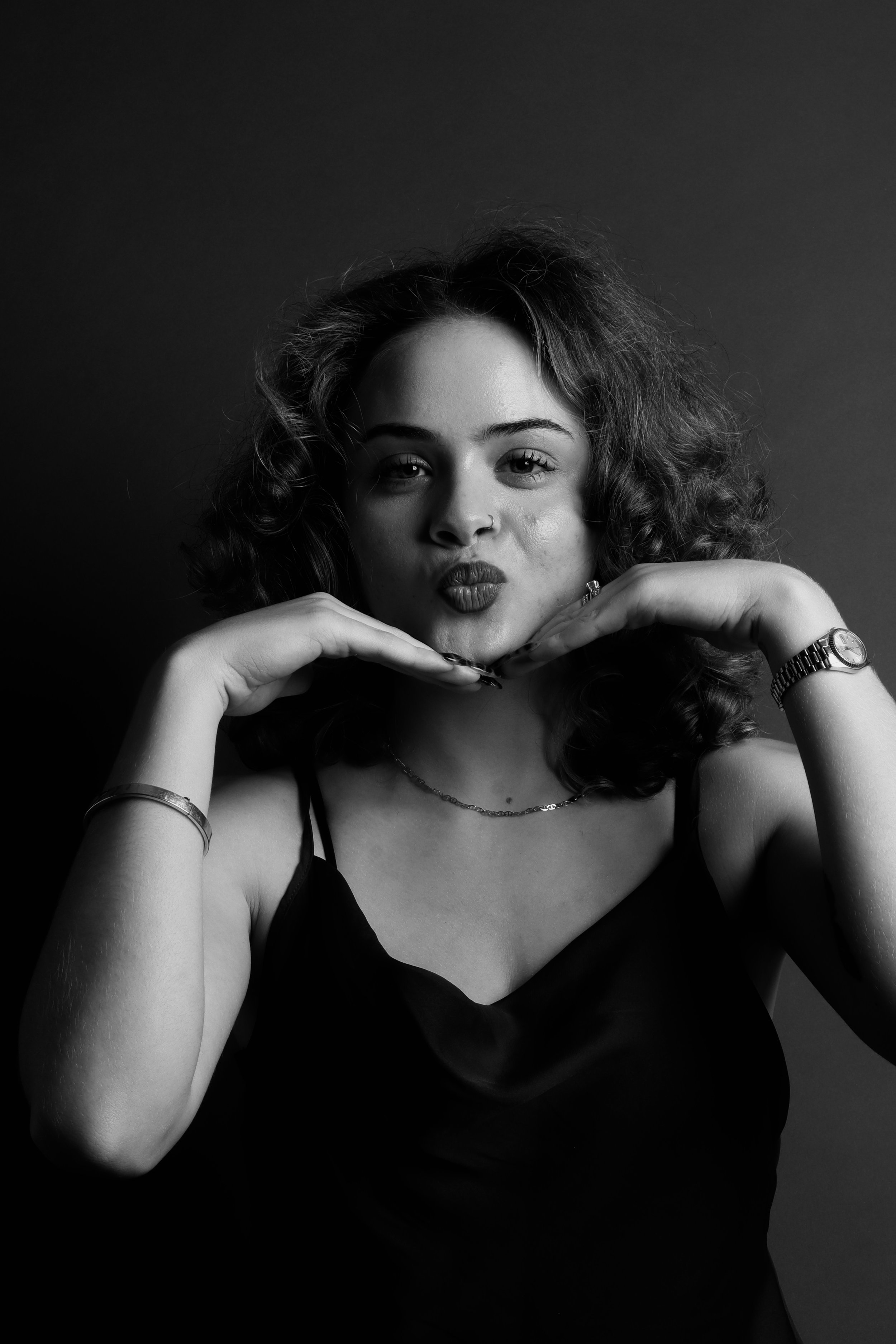 Black and white portrait of a young woman with curly hair, making a playful face with her lips puckered and hands under her chin, wearing a watch and jewelry.
