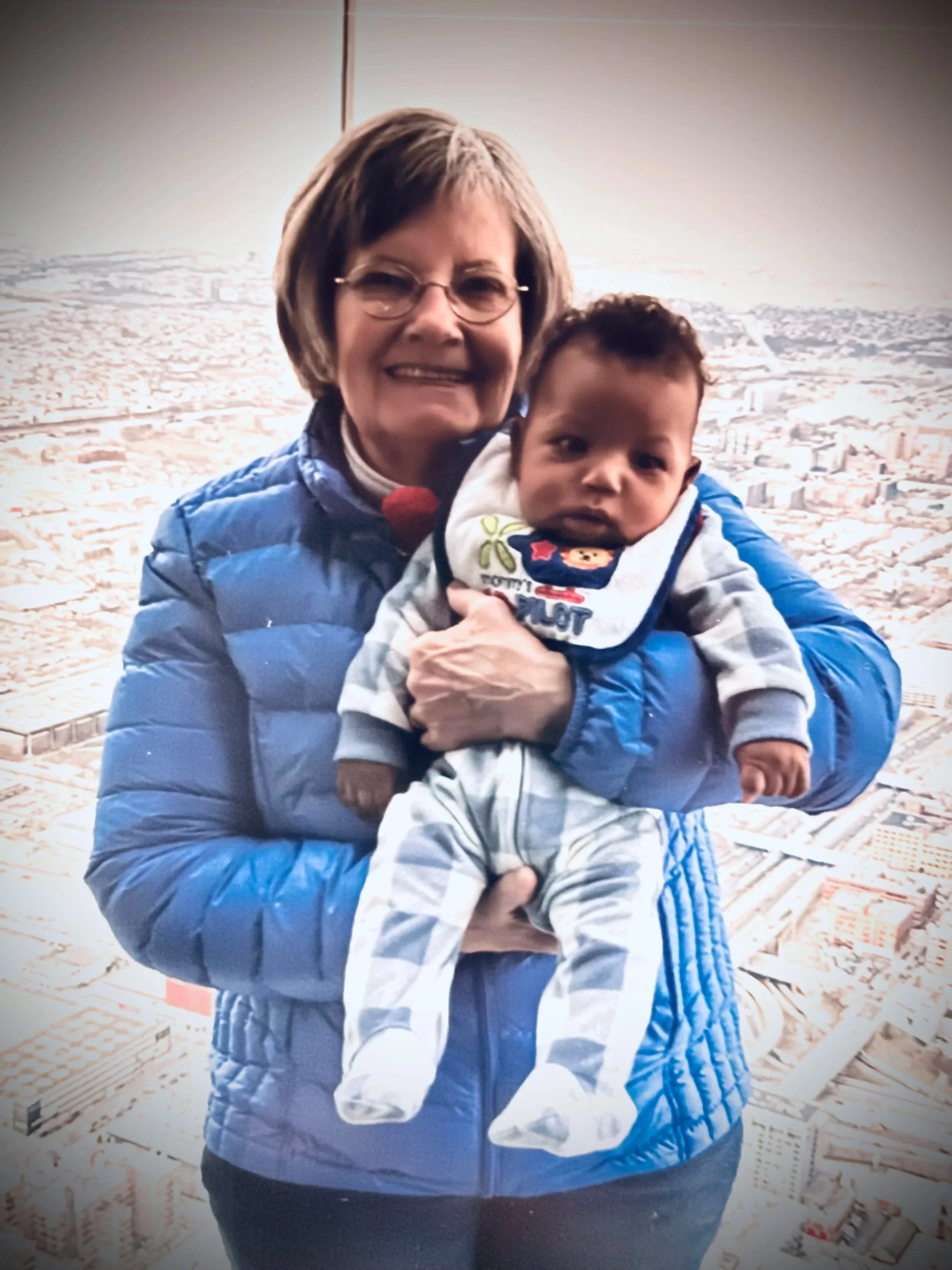 A woman with short brown hair, glasses, and wearing a blue puffer jacket, holding a baby in front of a cityscape with buildings and a hazy sky.