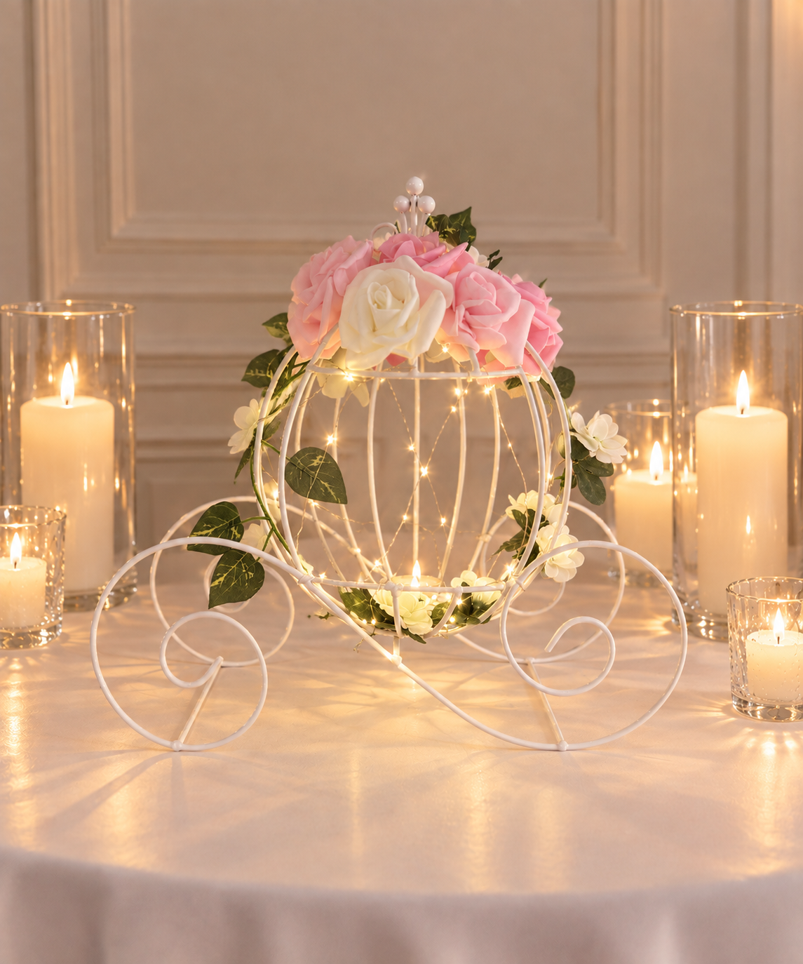 A decorative centerpiece on a table featuring a white metal carriage with pink and white flowers, surrounded by lit candles in glass holders, creating a warm ambiance.