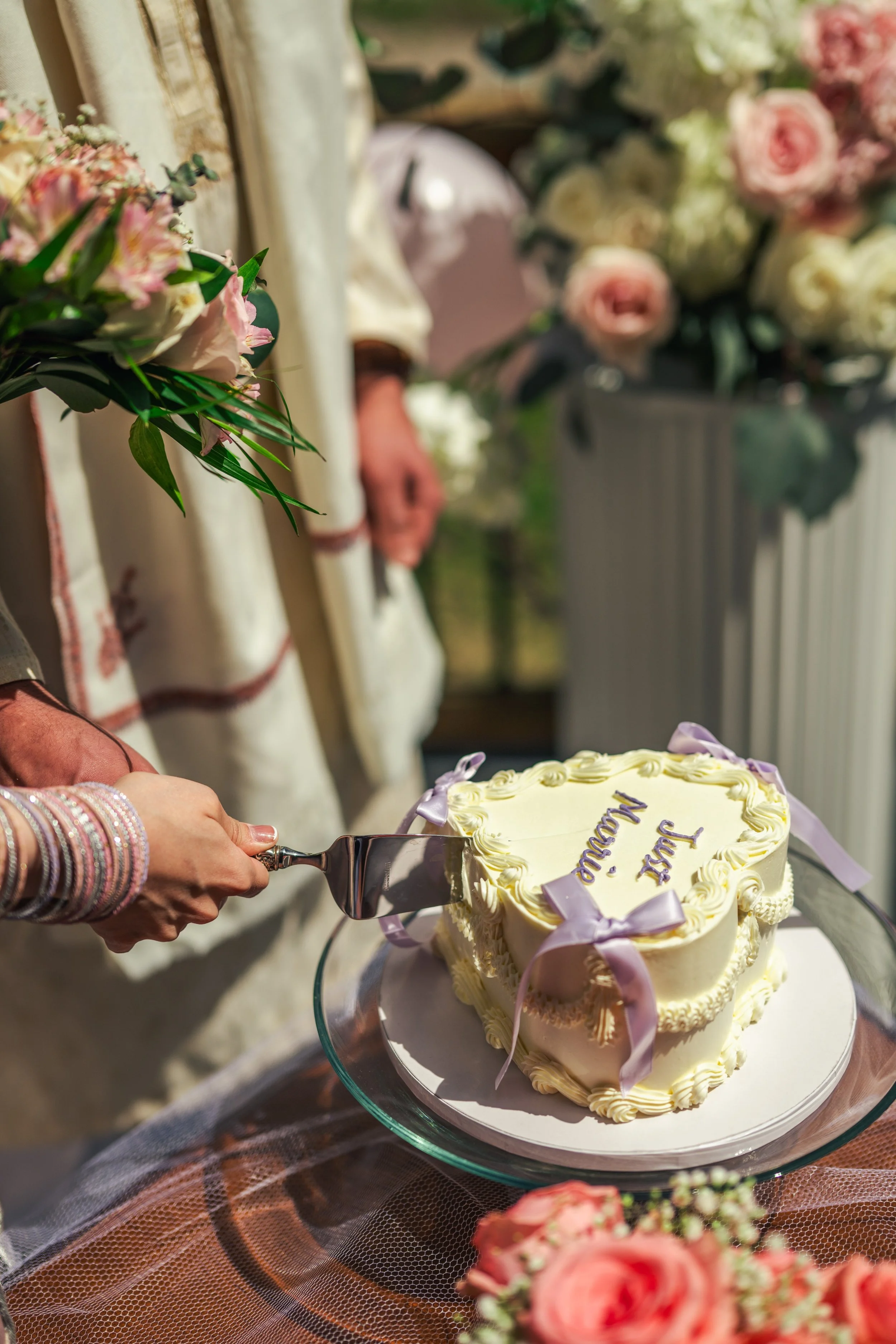A person in bracelets is cutting a white wedding cake with purple ribbons and writing, surrounded by pink and white flowers on a table covered with a brown mesh cloth.