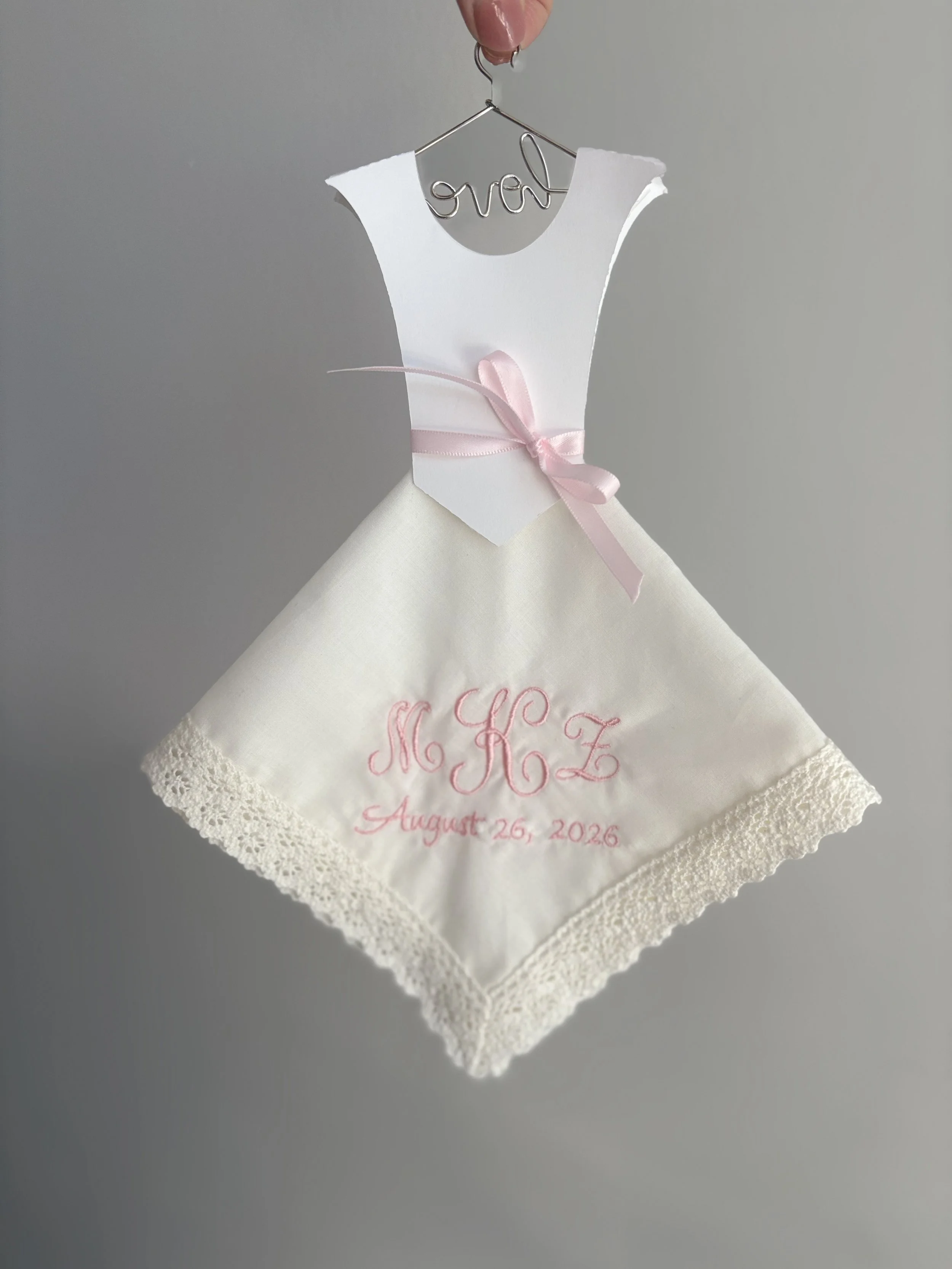 A small white dress with pink embroidered initials and date, hanging on a wire hanger with the word 'love' written in wire at the top.