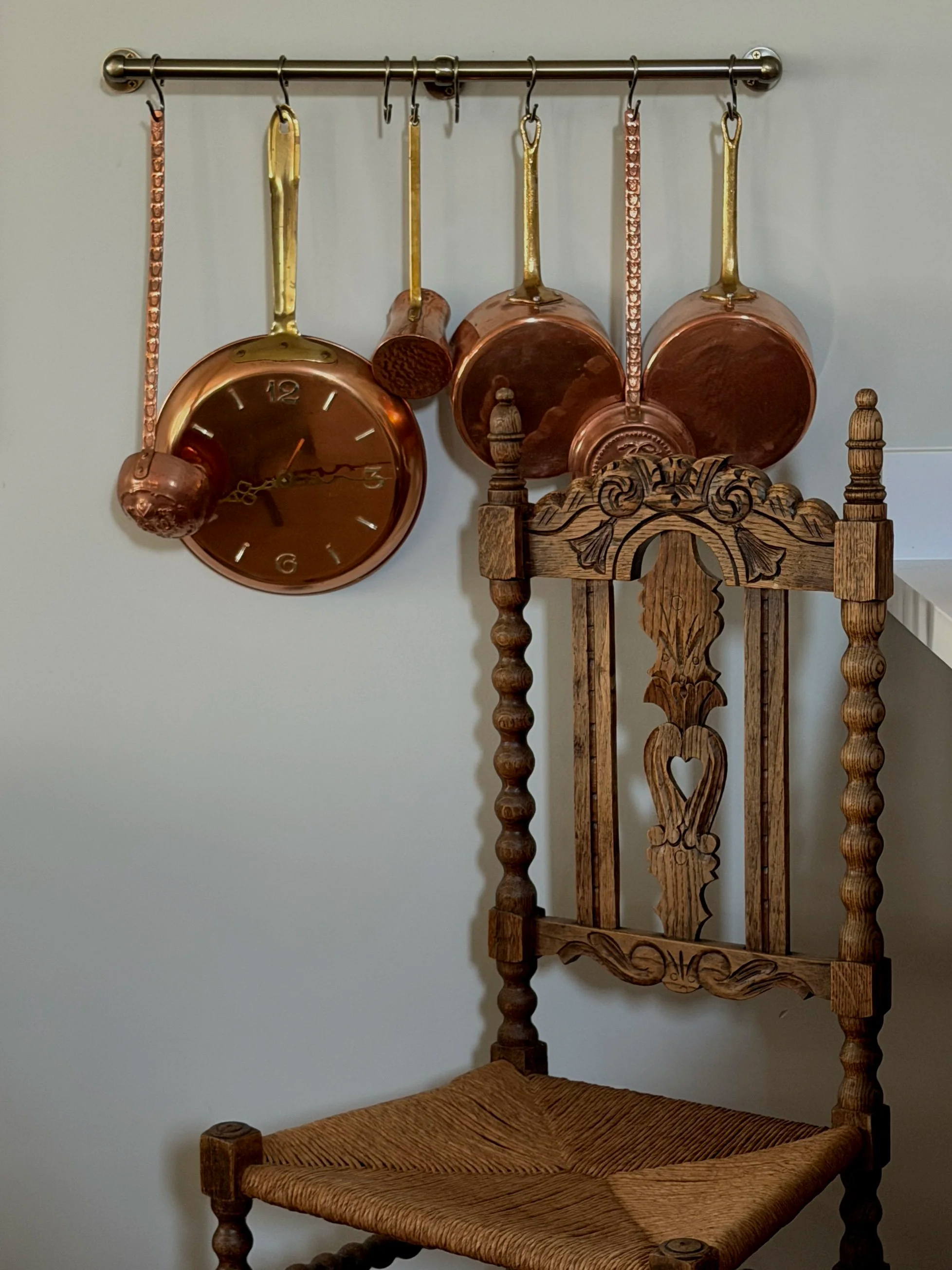 A wall-mounted metal rod with four hanging copper and gold kitchen utensils and a clock, alongside an ornate wooden chair with carved details and a woven seat.
