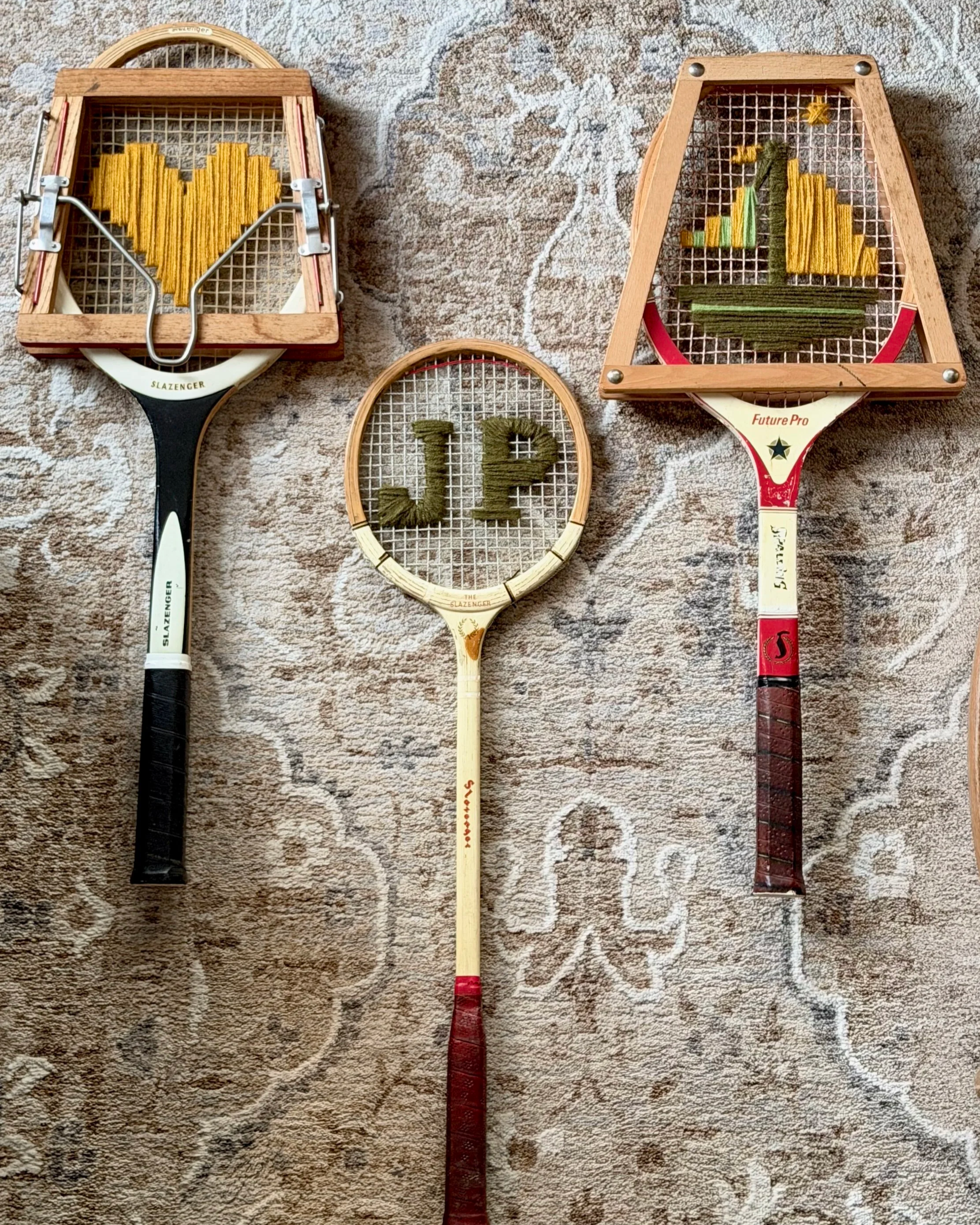 Three vintage tennis rackets on a patterned rug, each with a string art design on the racket heads. The left racket has a yellow heart, the middle has green letters 'L' and 'P', and the right has a yellow and green square shape with a yellow star on top.
