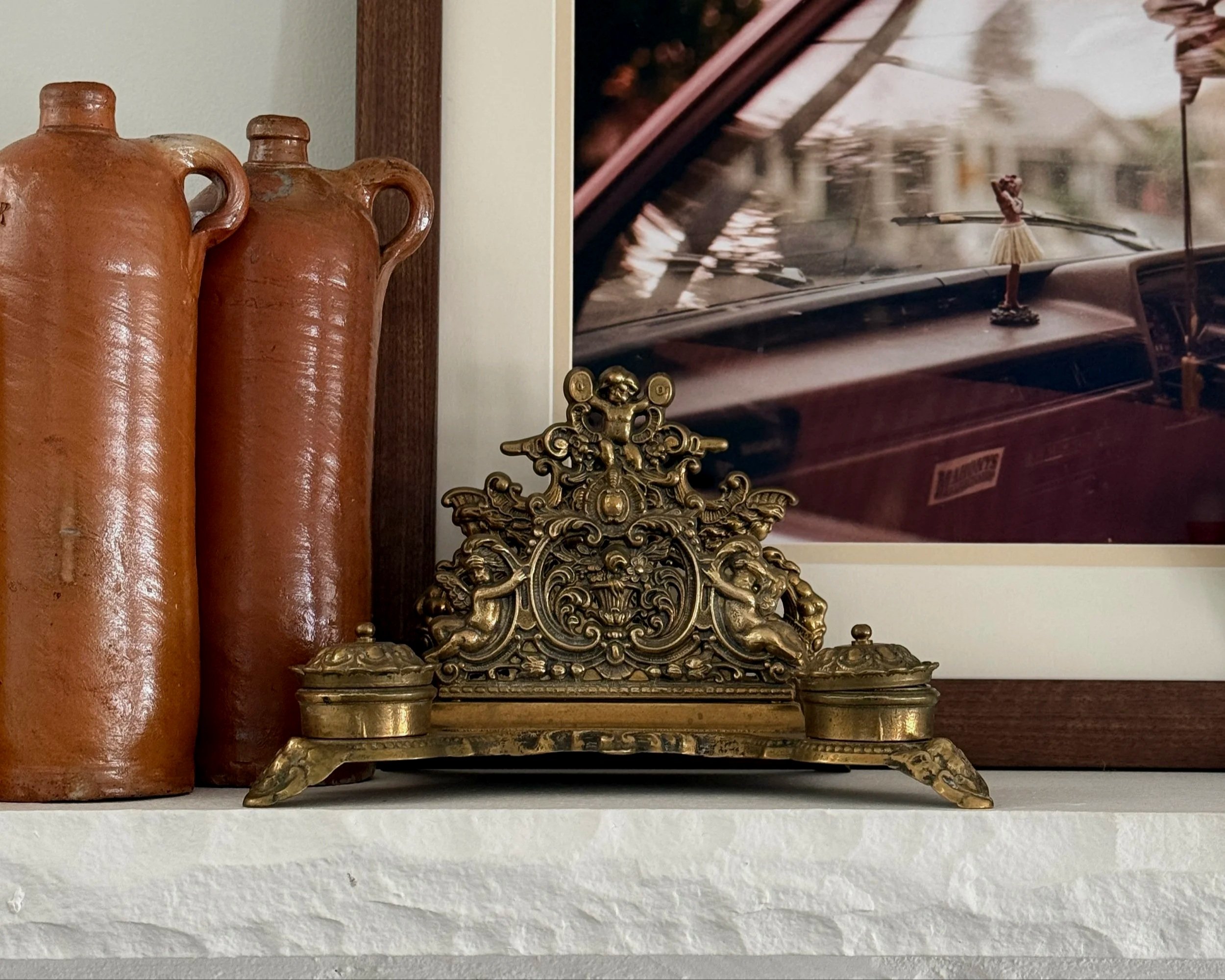 Decorative ornate brass or bronze altar piece placed on a white shelf, with two terracotta vases behind it and a framed artwork with motion blur of rainfall in the background.