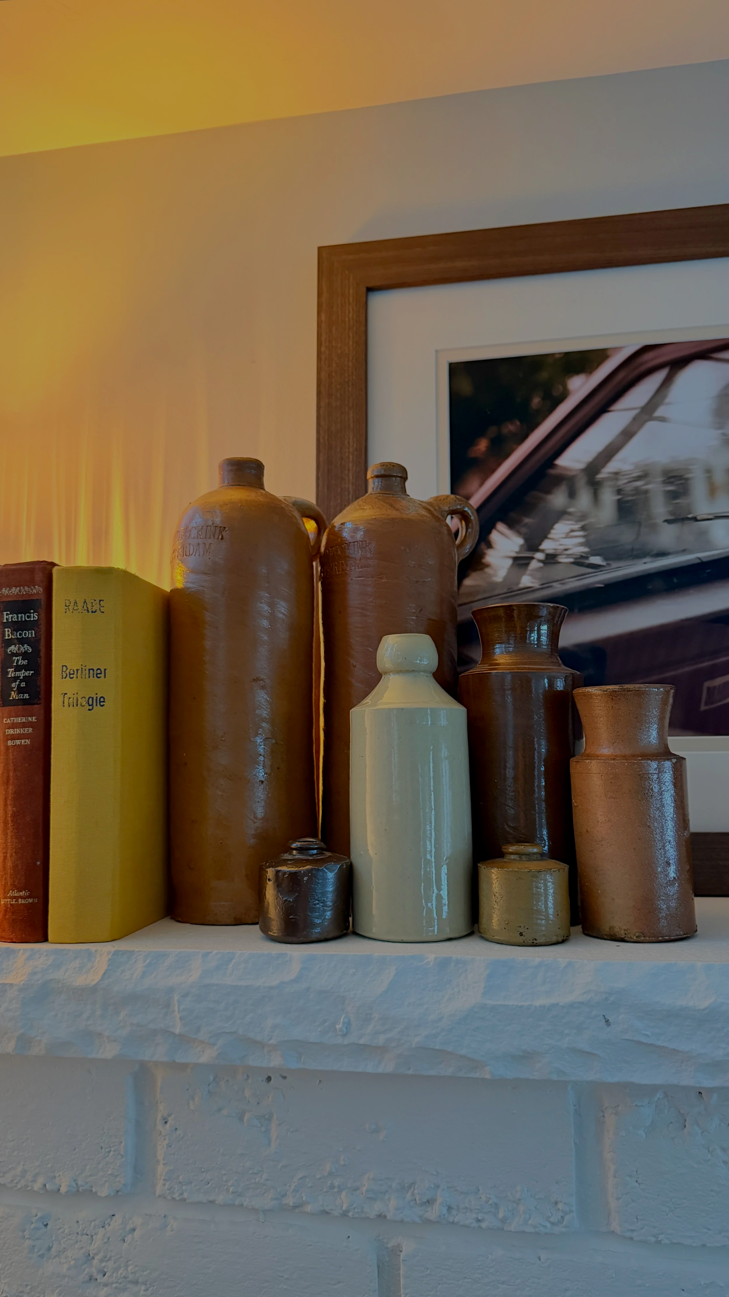 A collection of various-sized ceramic bottles and a book titled "Race Berliner Trilogie" on a white brick shelf, with a framed photograph of a car interior in the background.