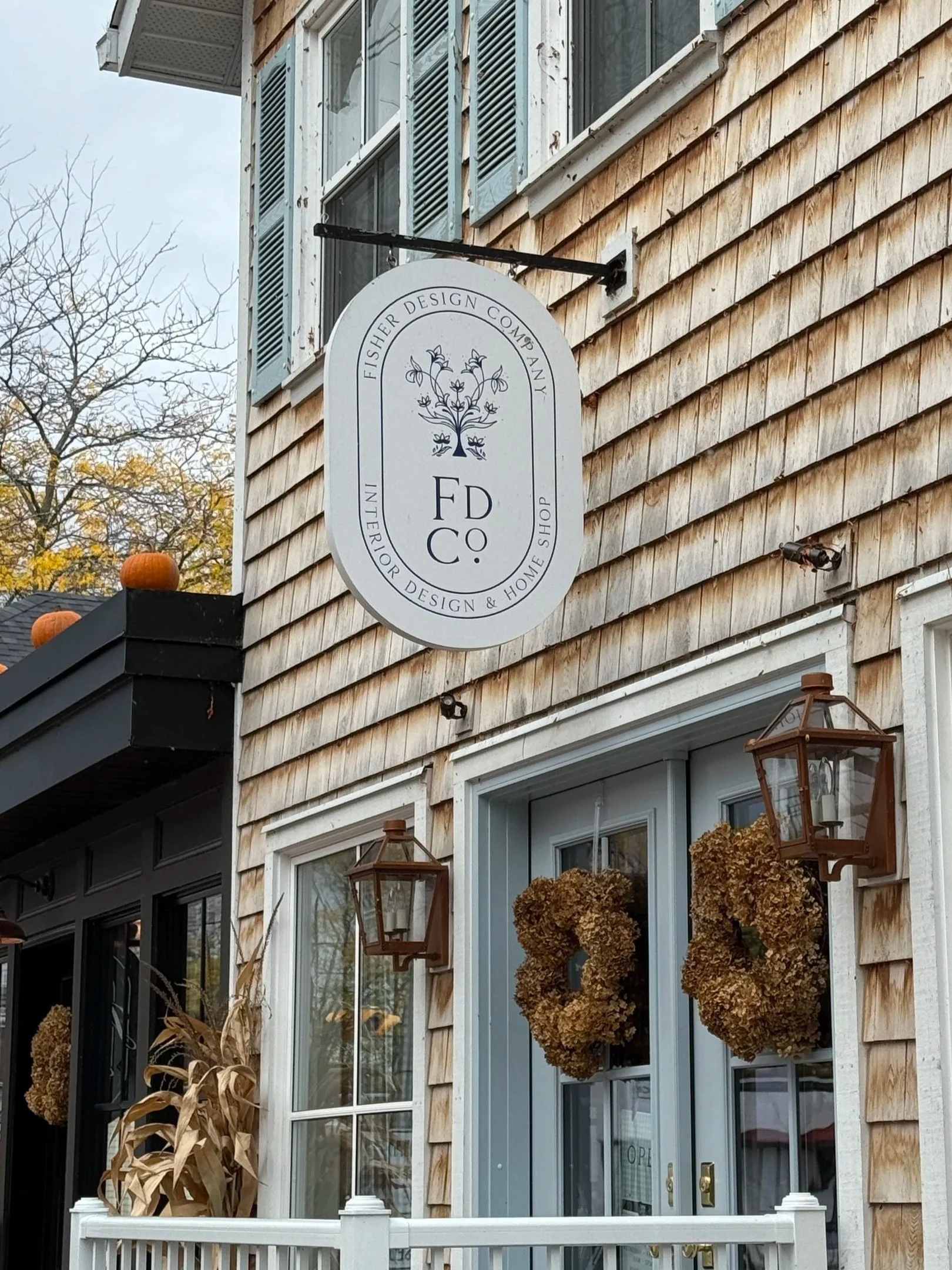 Exterior of a building with wood siding, large window, and a sign for Fisherer Design Company. The sign mentions interior design and home shop. There are plants and outdoor lanterns near the entryway.
