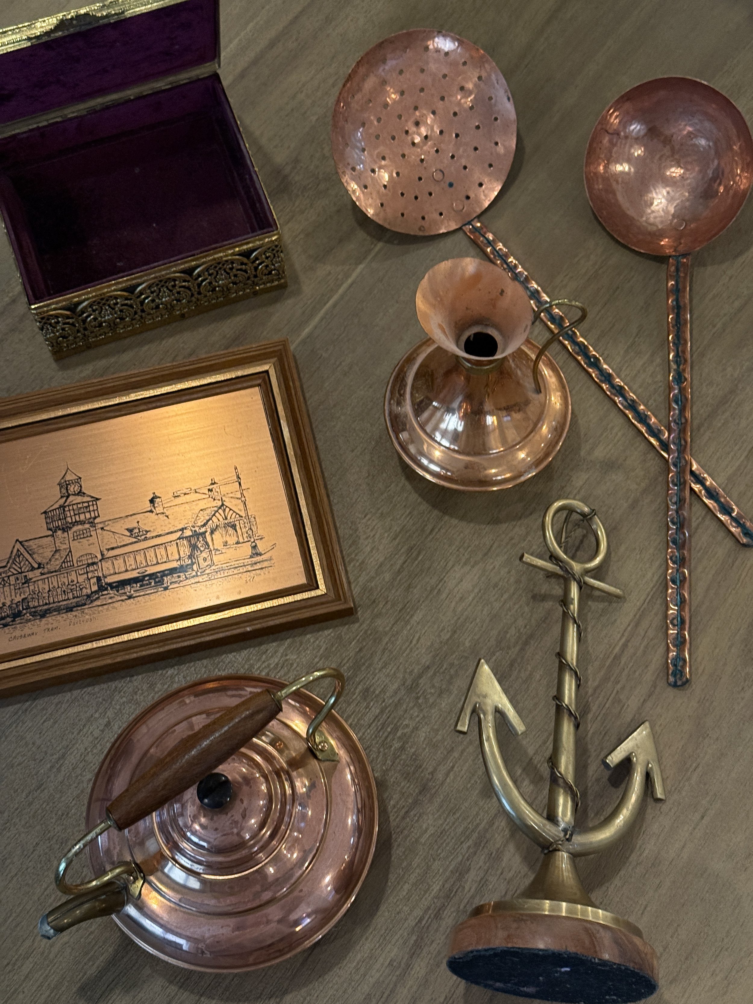 Decorative copper and brass objects, including a finished copper watering can, a box with purple lining, framed artwork, and various metallic decorative items on a wooden surface.
