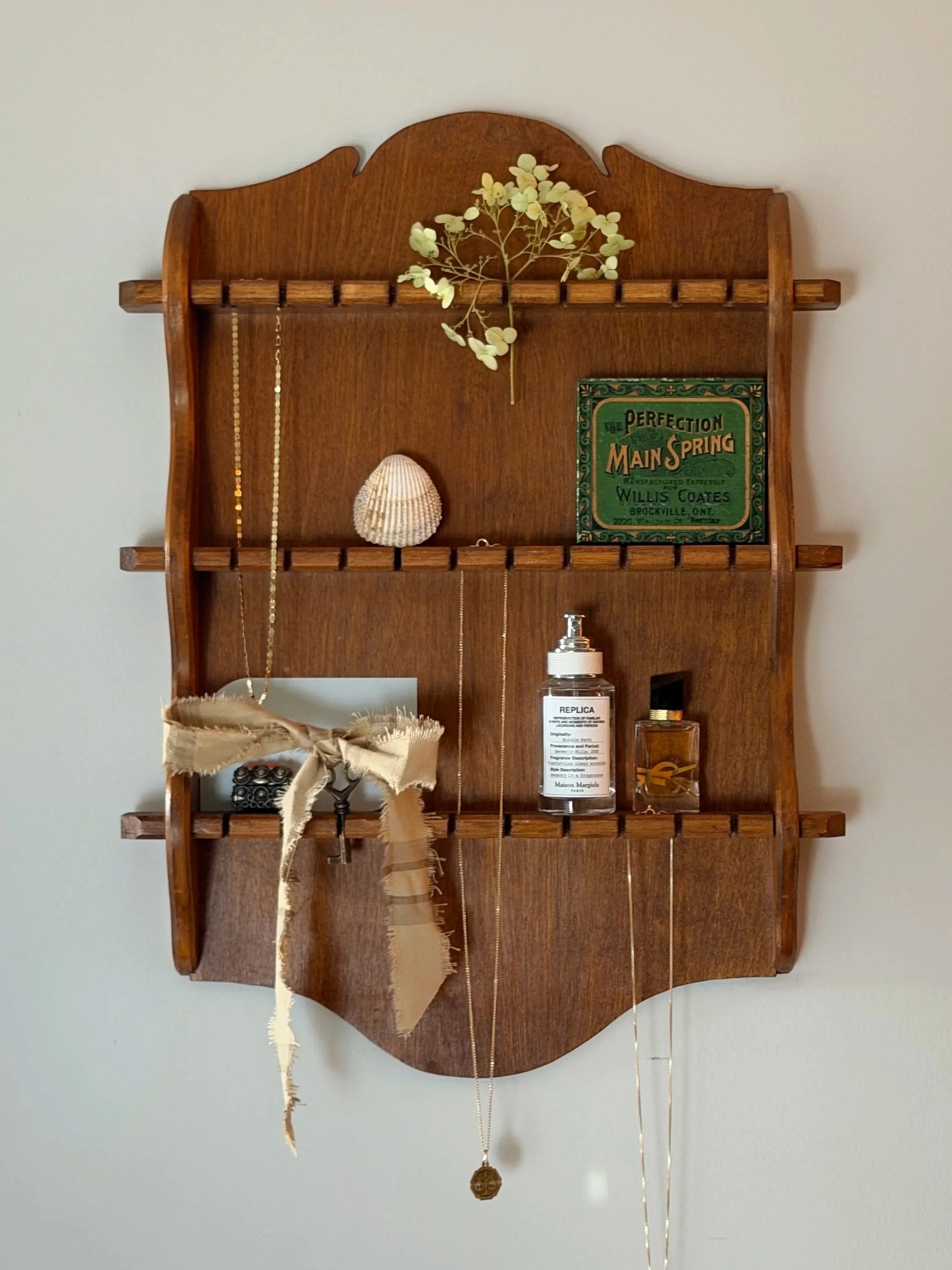 Wall-mounted wooden shelf with decorative items including a dried flower, a seashell, a green vintage sign, a small glass jar with a label, and a hanging chain with a round pendant.