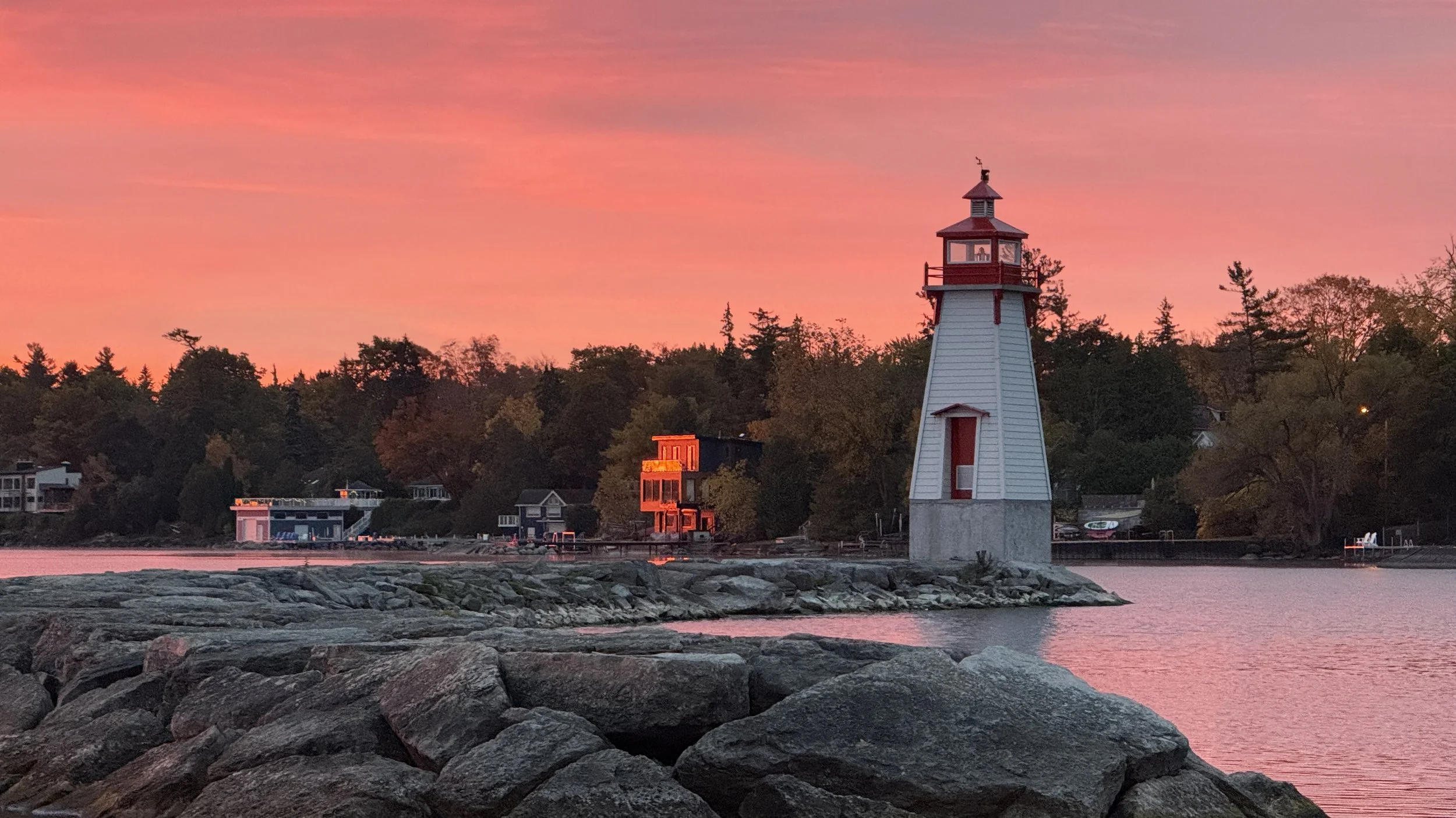 A lighthouse on a rocky shoreline during a colorful sunset, with trees and houses in the background.