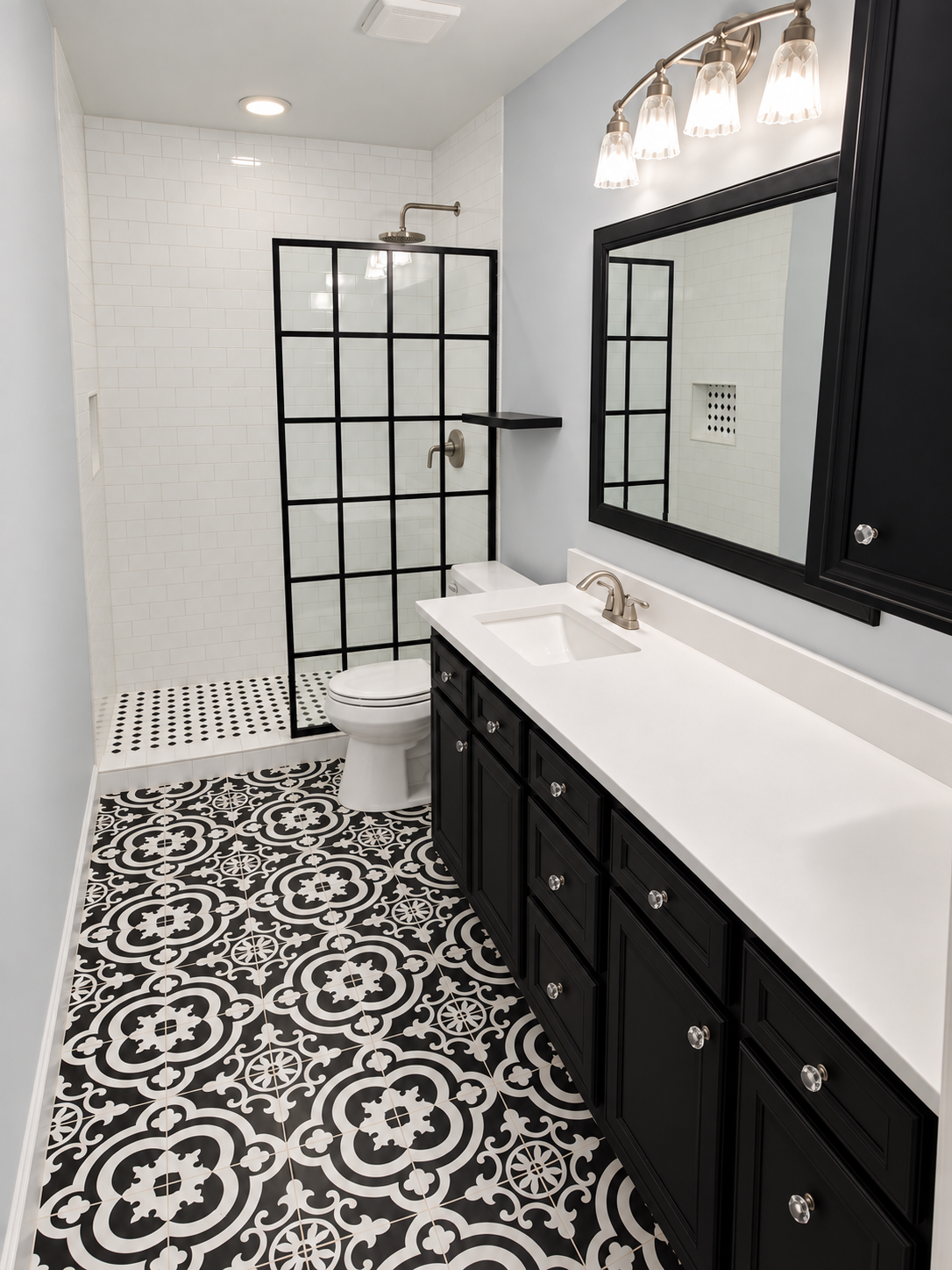 Modern bathroom with black and white patterned floor tiles, black vanity with silver knobs, white countertop, large mirror, glass light fixture, and a walk-in shower with black framing and white tiles.