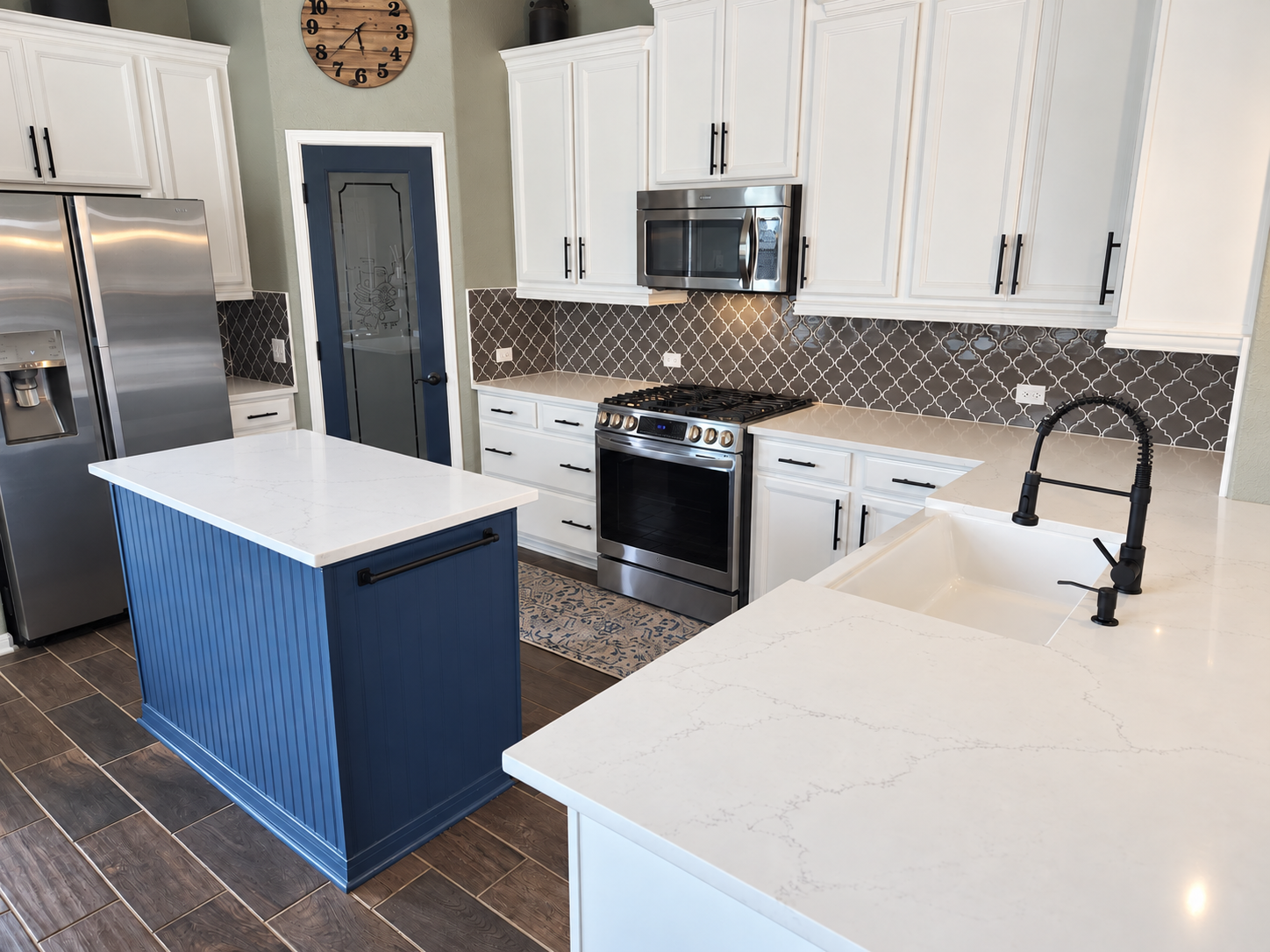 Modern kitchen with white cabinets, a stainless steel refrigerator, a built-in microwave above a stove, a blue kitchen island with a white marble top, a white countertop and sink, dark wood flooring, and a wall clock.