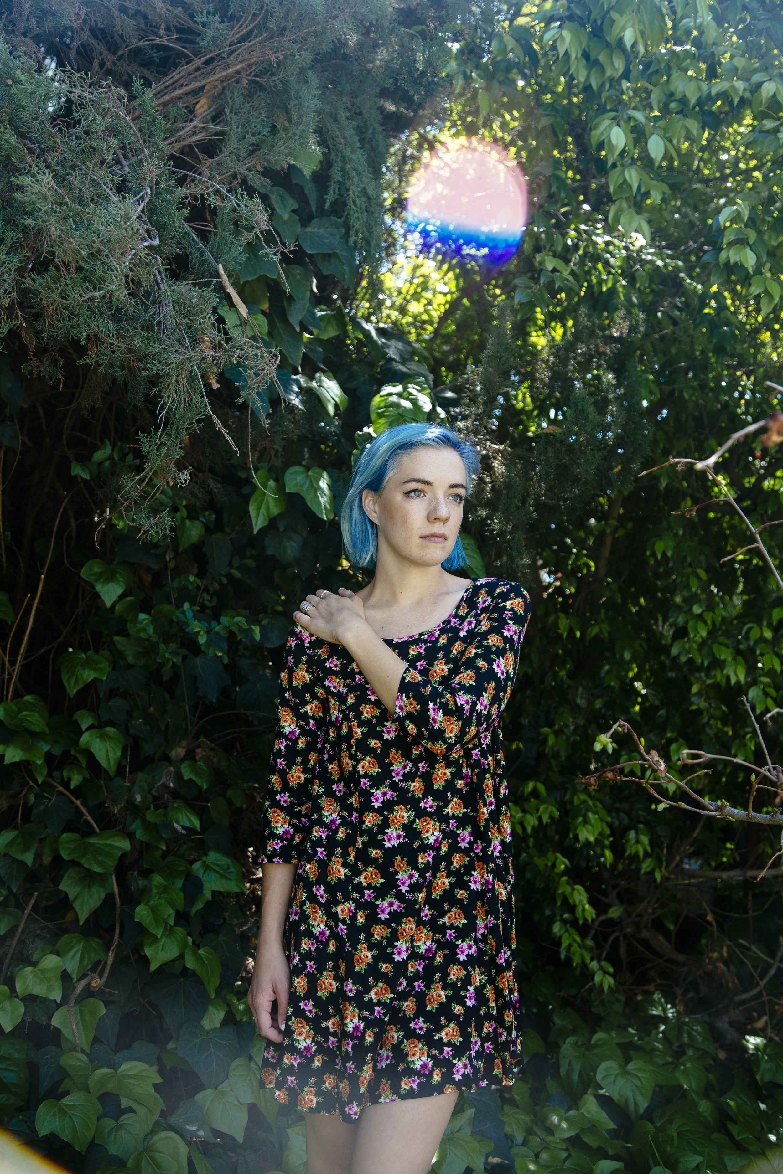 A young woman with blue hair wearing a black floral dress stands amidst green foliage and vines, with sunlight filtering through the trees. She has a contemplative expression.