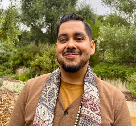 A man smiling outdoors with trees and bushes in the background, wearing a brown jacket and a patterned scarf.