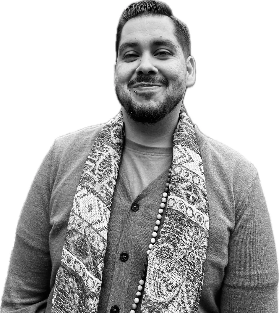 A smiling man with a beard and mustache, wearing a cardigan, a T-shirt, a patterned scarf, and a beaded necklace.