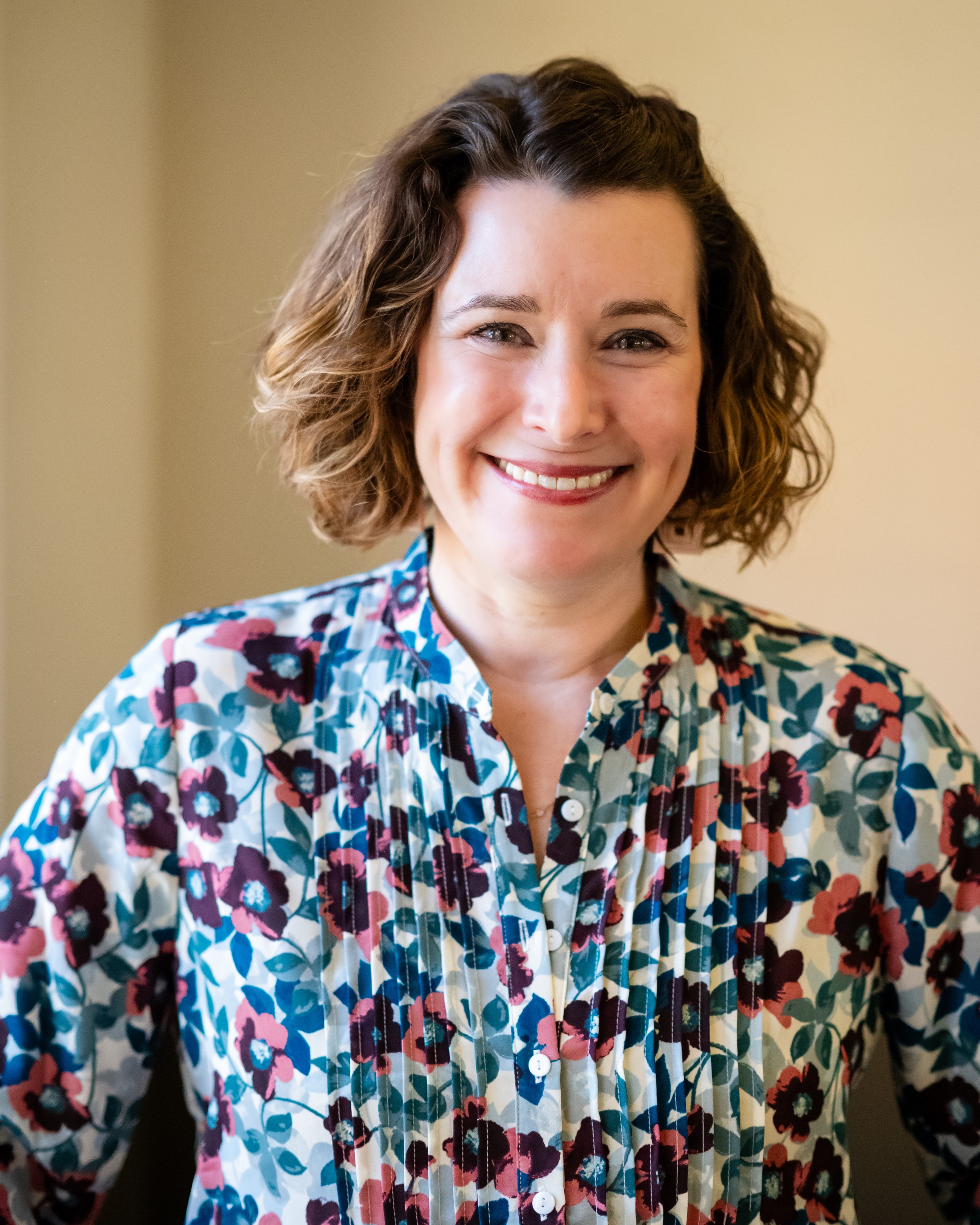 A woman with shoulder-length wavy brown hair, smiling with visible teeth, wearing a colorful floral patterned blouse with a white background decorated with blue, purple, red, and green flowers. The background is beige and out of focus.
